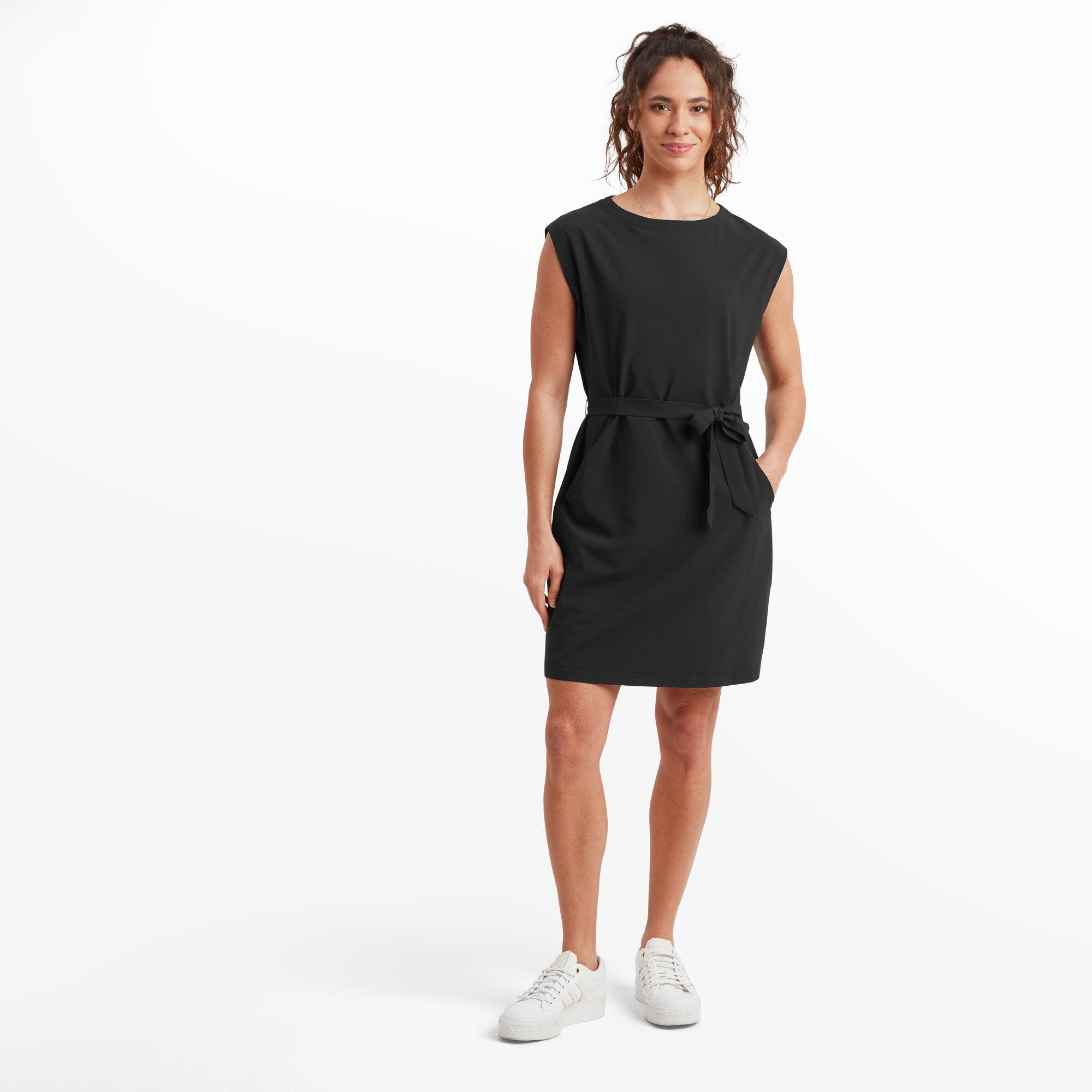 A woman is wearing a sleek Sherpa Adventure Gear Sajilo Travel Dress in Black that features a slightly structured design. The dress is cinched at the waist with a matching fabric tie, creating a flattering silhouette. She pairs the outfit with white trainers, giving it a casual yet polished look. She has a relaxed pose with one hand on her hip and the other hanging by her side, exuding confidence and style.