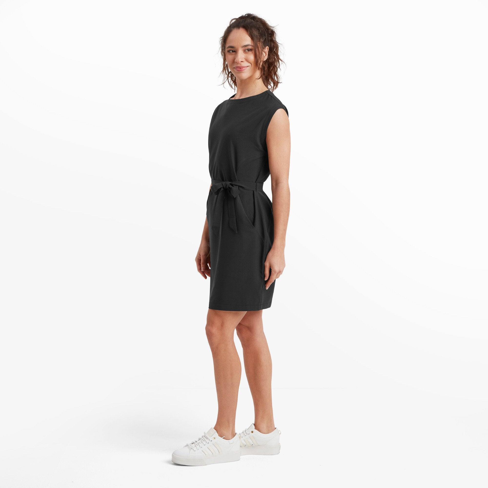 The model is captured from a slight side angle, highlighting the tailored fit of the Sherpa Adventure Gear Sajilo Travel Dress in Black. The fabric tie at the waist is clearly visible, emphasising the adjustable nature of the fit. Her posture and smile give a casual and approachable vibe. The outfit remains paired with the same white trainers, maintaining a cohesive and comfortable aesthetic.