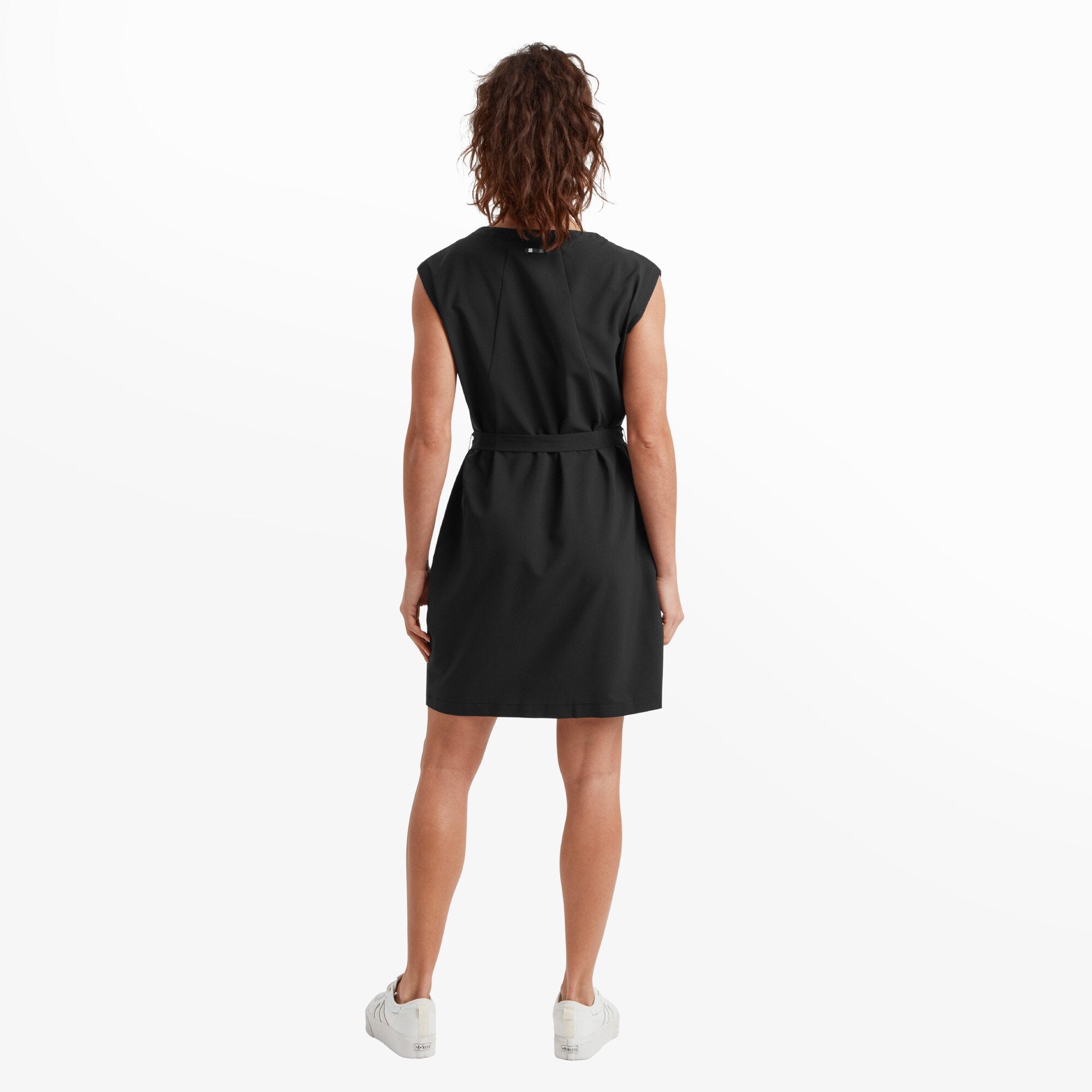 This image shows the back of the Sherpa Adventure Gear Sajilo Travel Dress in Black, with its clean lines and simple design. The waist tie continues around the back, adding to the structured look. The model's wavy hair frames her back as she stands with relaxed posture, and the black dress contrasts nicely with the white trainers for a modern, minimalist style.