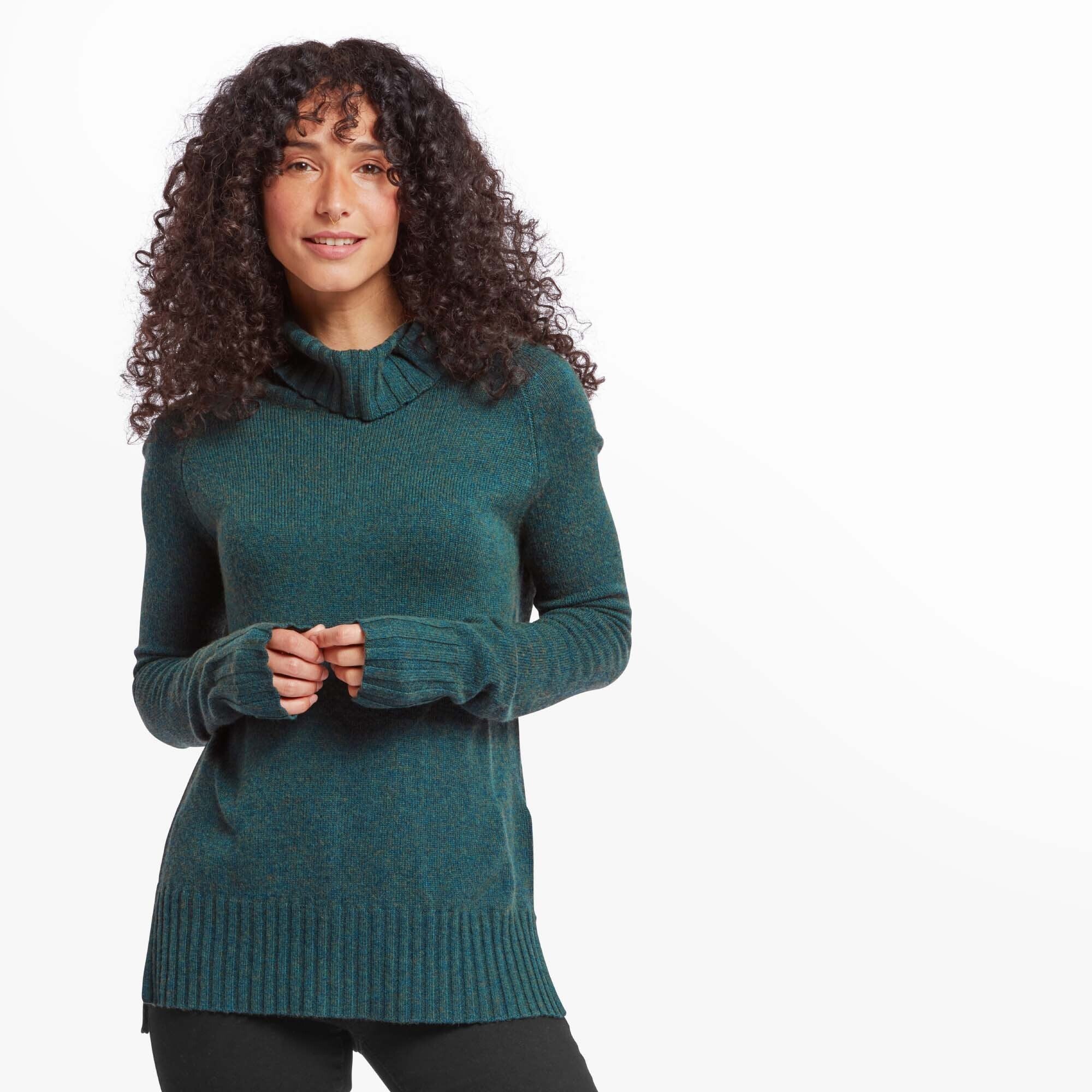 A woman with curly dark hair and light brown skin is wearing a Sherpa Adventure Gear Saleena Eco Roll Neck in Green with long sleeves and a relaxed fit. She is standing against a plain white background, looking directly at the camera with a slight smile. The sweater has a high, fold-over neckline and extended ribbed cuffs with thumbholes.