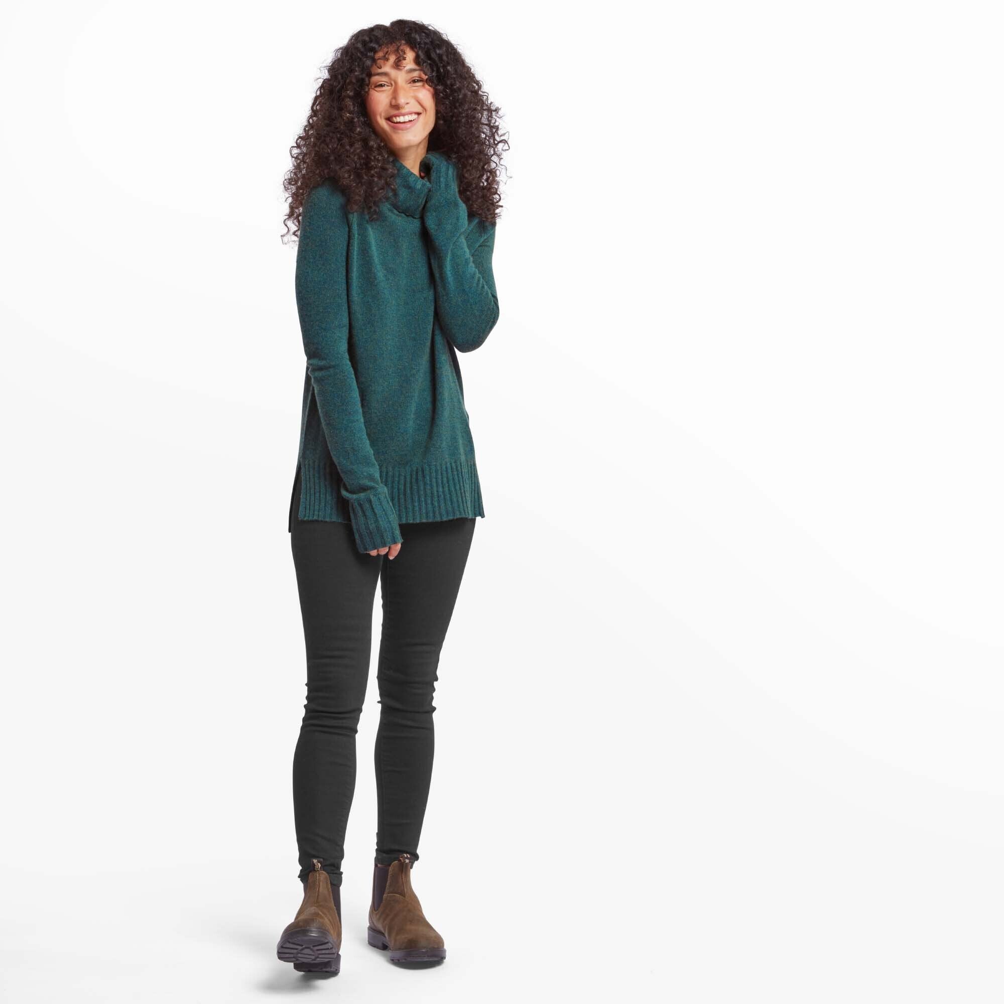 The same woman is posing in a full-body shot, wearing the Sherpa Adventure Gear Saleena Eco Roll Neck in Green paired with black skinny jeans and brown ankle boots. She is standing with her weight shifted to one side, smiling brightly, with her right hand gently pulling at the neckline of the sweater. The sweater drapes over her hips, with a ribbed hem.