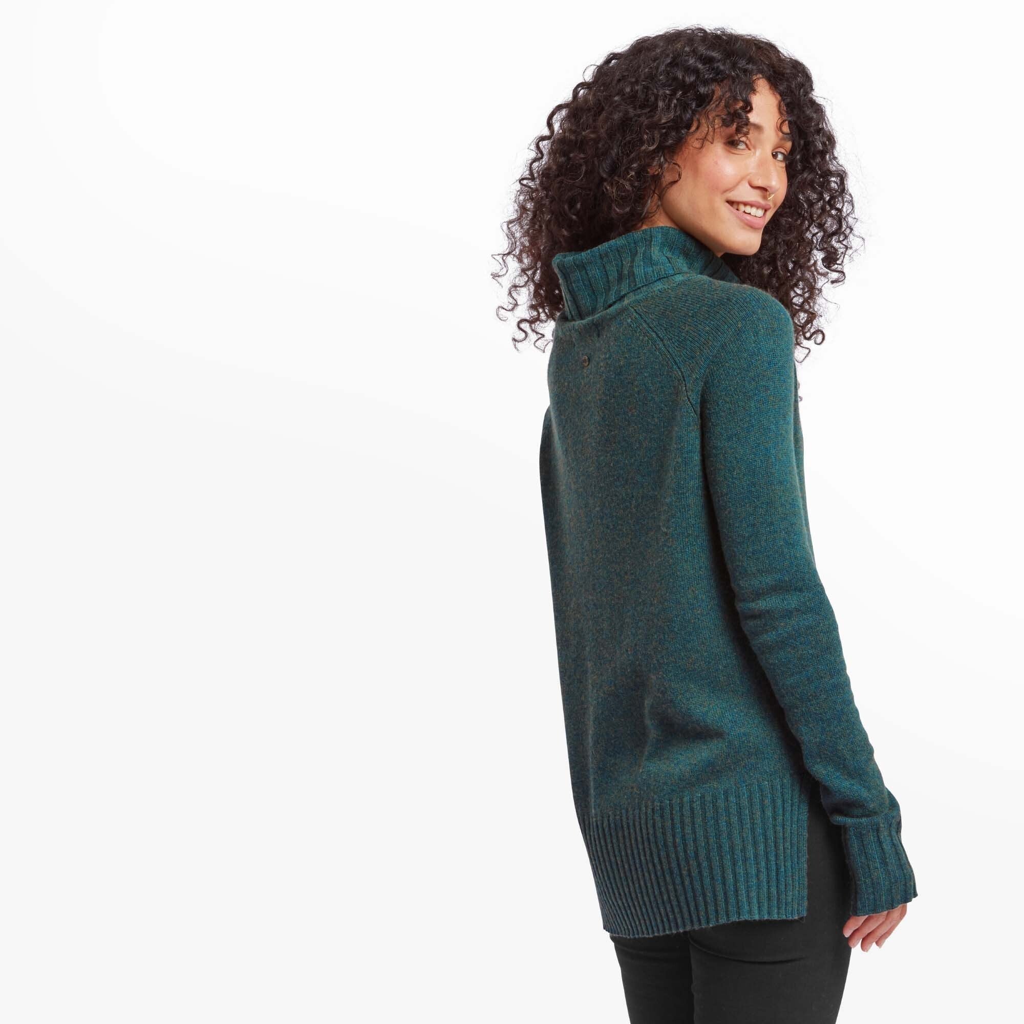 A side-angle image of the woman showing the back of the Sherpa Adventure Gear Saleena Eco Roll Neck in Green. She is turning her head toward the camera with a playful smile. The sweater has a slightly longer back hem and visible ribbed detailing along the edges.