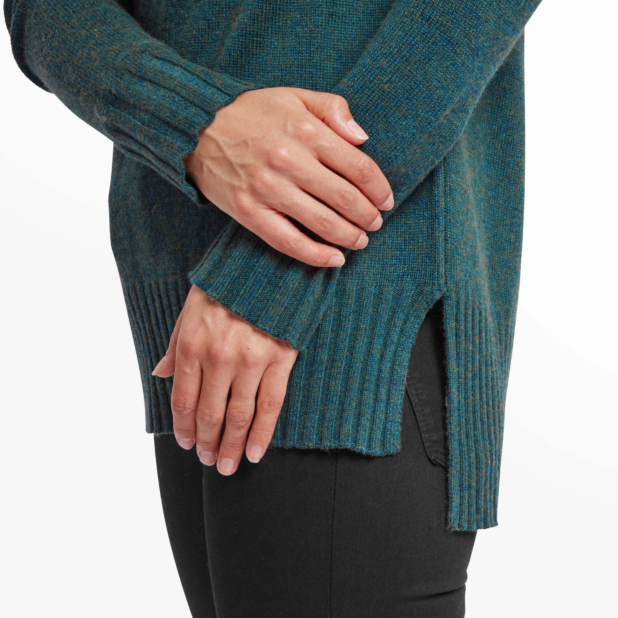 A close-up shot of the woman's hands, focusing on the ribbed texture of the Sherpa Adventure Gear Saleena Eco Roll Neck in Green's cuff and hem. The thumbhole in the extended cuff is visible as she gently rests her hands on each other. The sweater’s side slit and knit pattern detail are evident in the close-up.