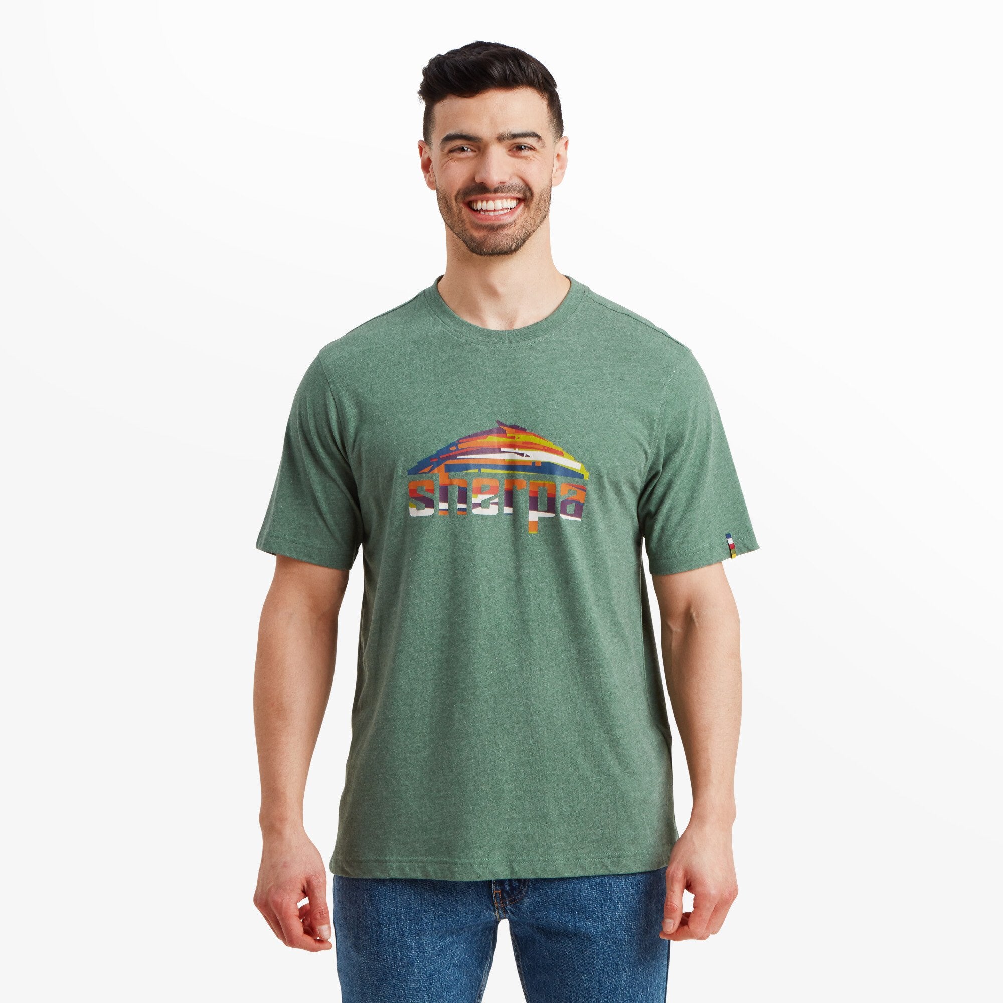 A male model with a fit physique, short dark hair, and a well-groomed beard is wearing a Sherpa Adventure Gear Sherpa Mountain T-Shirt in Green with a colorful Sherpa mountain logo on the chest. He is smiling warmly and looking directly at the camera. The T-shirt is made from a heathered fabric, has a relaxed fit, and falls just below the waist. The model pairs it with classic blue jeans, standing against a plain white background.