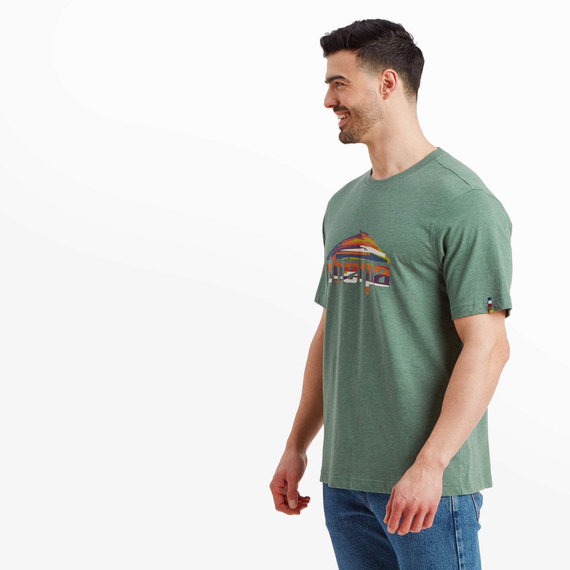 The same male model is captured in a three-quarter side view, looking to his left. The Sherpa Adventure Gear Sherpa Mountain T-Shirt in Green has a straight-cut hem, falling naturally over his jeans. The vibrant Sherpa mountain logo is still visible from the side. The model maintains a relaxed posture with his arms slightly away from his body, showing the comfortable fit of the fabric.