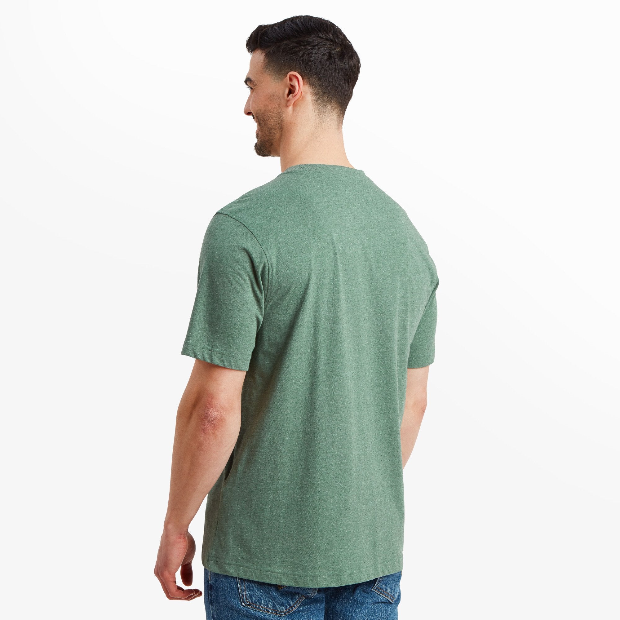 The model is now turned away from the camera, showing the back of the Sherpa Adventure Gear Sherpa Mountain T-Shirt in Green. The design is simple with no visible branding on the back. The T-shirt fits loosely over his shoulders and upper back, draping naturally. The sleeves fall just above the elbow, and the hemline reaches past the waistline of his jeans.