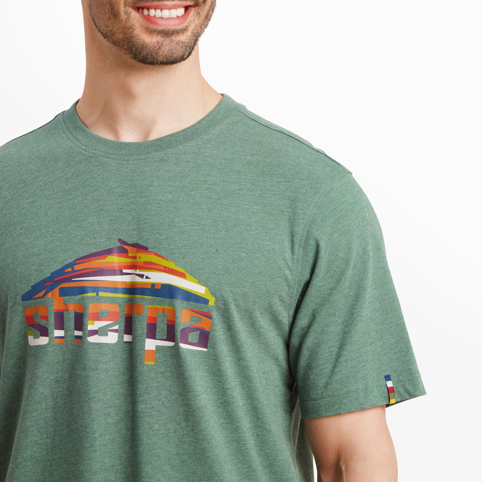 A zoomed-in view of the Sherpa logo on the chest of the Sherpa Adventure Gear Sherpa Mountain T-Shirt in Green. The logo is a stylized mountain illustration in shades of red, blue, yellow, and orange, with the word "Sherpa" written in bold, multicolored capital letters below. The texture of the fabric is visible, showing a heathered blend. A small, multicolored striped tag is sewn onto the left sleeve.