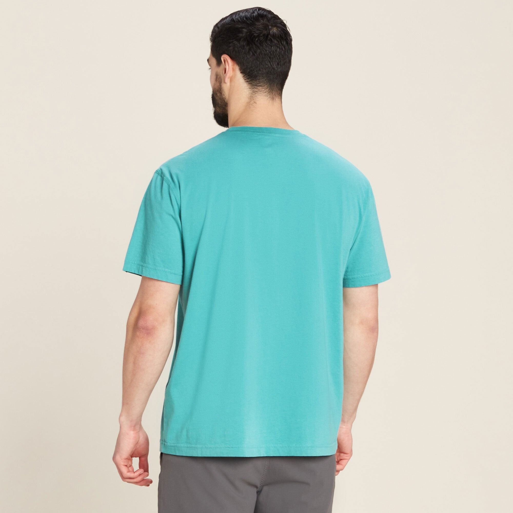 The same male model is shown from the back, wearing the Sherpa Adventure Gear Sokaa T-Shirt in Blue. The shirt has a clean, plain back with no visible logos or designs. It has a standard length and falls just below the waist. The model's dark grey pants are also visible. The background is a plain neutral beige.