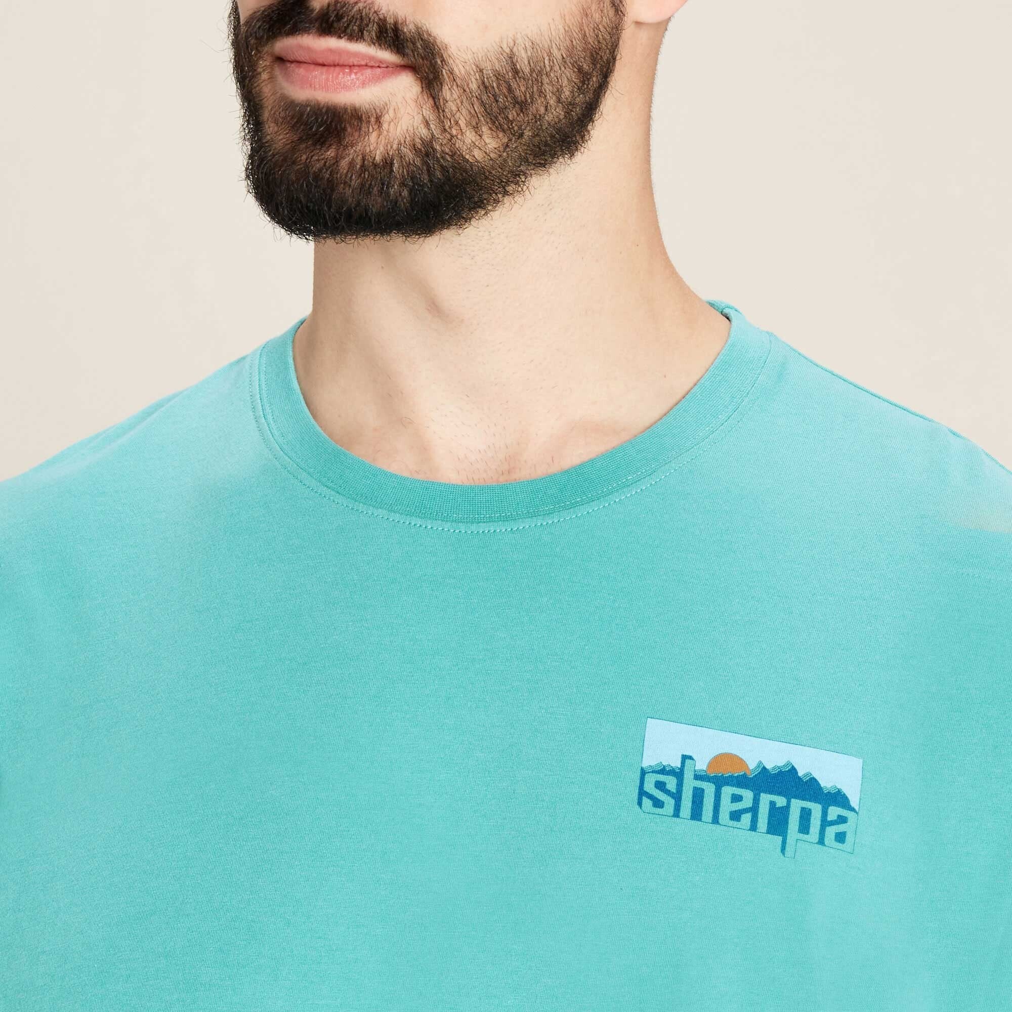 A close-up image of the upper left chest area of the Sherpa Adventure Gear Sokaa T-Shirt in Blue. The logo is a small rectangular patch with the word "Sherpa" written in bold lowercase letters. Above the text is a stylized graphic of blue mountains with a rising or setting sun in orange. The fabric appears to be soft cotton with a slight texture. The model's beard and lower face are partially visible in the upper part of the image. The background is neutral beige.