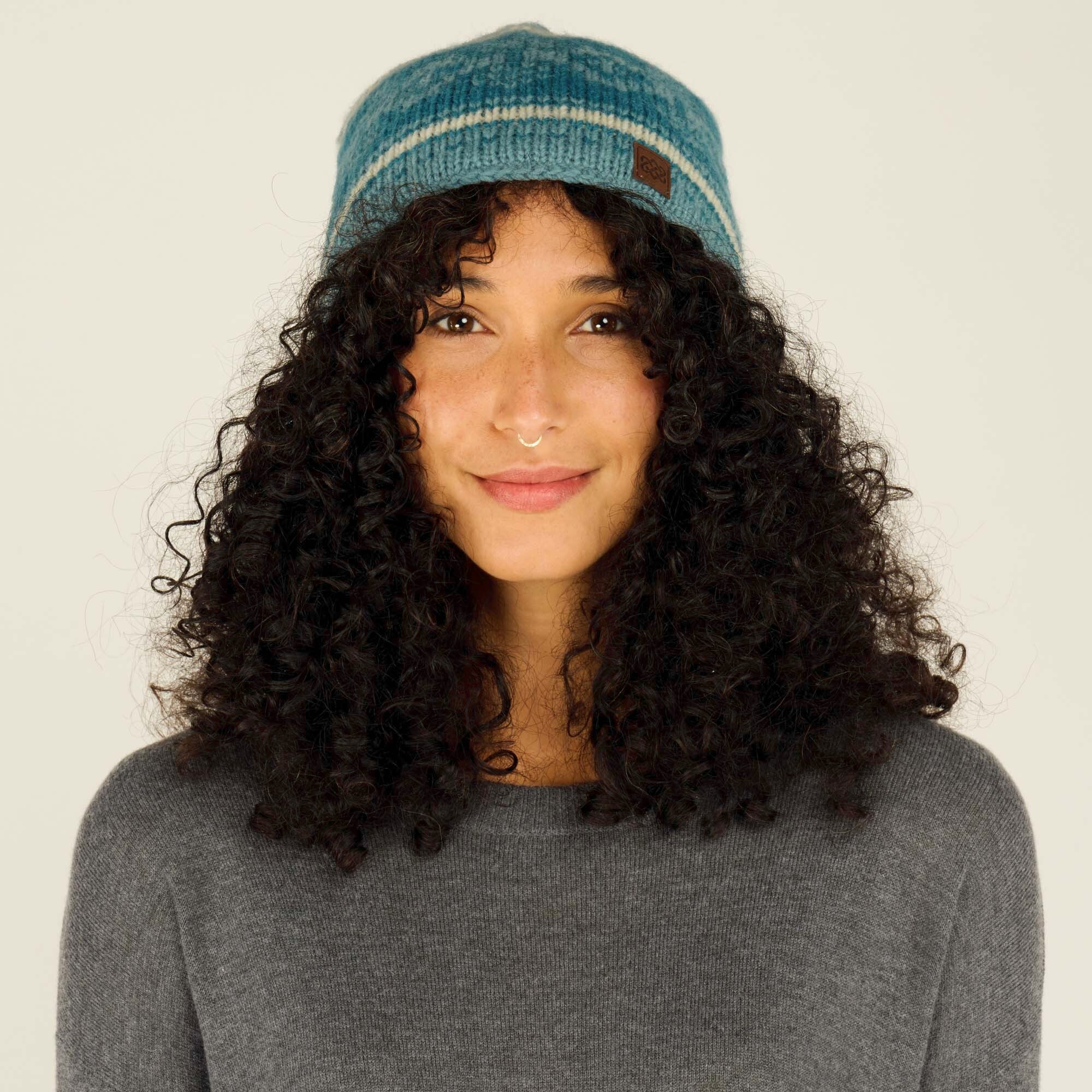 A close-up image of a woman with medium skin tone and dark curly hair wearing a Sherpa Adventure Gear Soni Hat in Green. The hat is made of soft knitted fabric with horizontal stripes in shades of blue and off-white. A small brown leather logo patch is sewn onto the lower left side of the hat. The woman is smiling gently, with her hair cascading down her shoulders. She is wearing a gray sweater, and her nose is adorned with a small gold septum ring. The background is a neutral beige color.