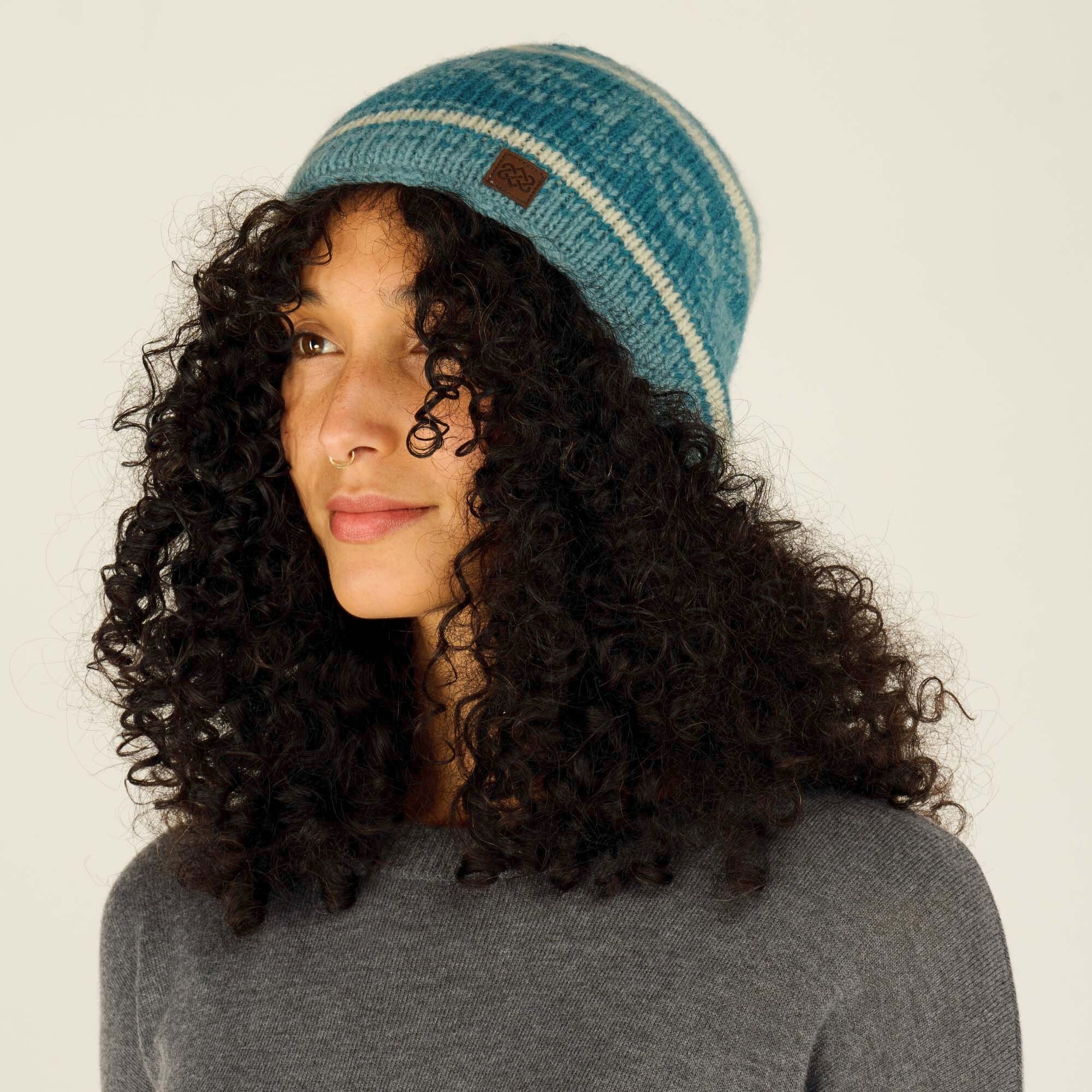 A side profile image of the same woman wearing the Sherpa Adventure Gear Soni Hat in Green. The hat’s horizontal striped pattern is clearly visible, with alternating bands of blue and off-white. The small brown leather logo patch is seen more clearly from this angle. Her curly hair frames her face, and her gold septum ring is visible. She is wearing the same gray sweater, and the background remains a neutral beige.