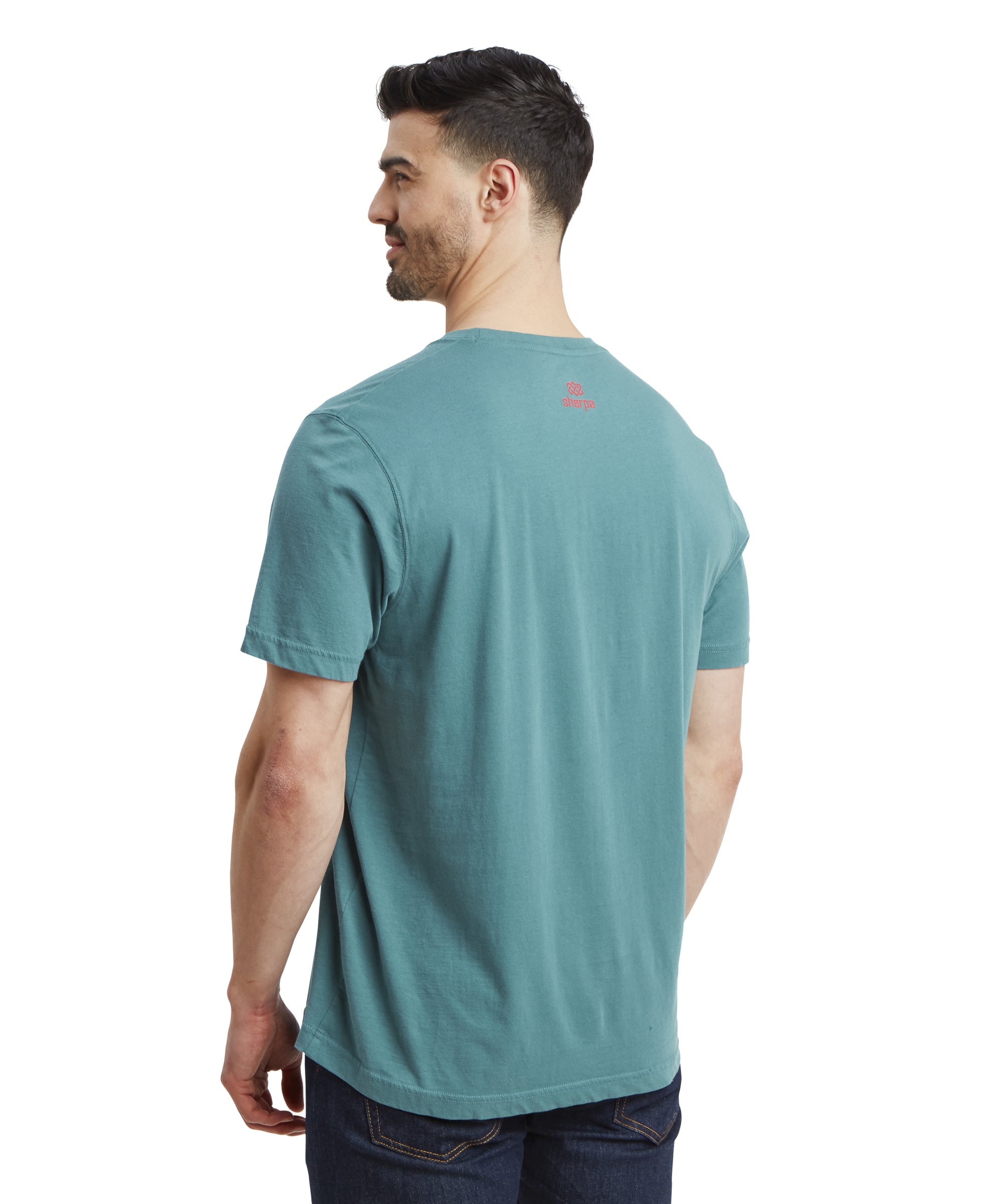 The back view of the man wearing the Sherpa Adventure Gear Tarcho T-Shirt in Blue, showing the Sherpa logo in red printed at the upper back of the shirt. He is wearing dark denim jeans, and the t-shirt hangs naturally over his waistline.