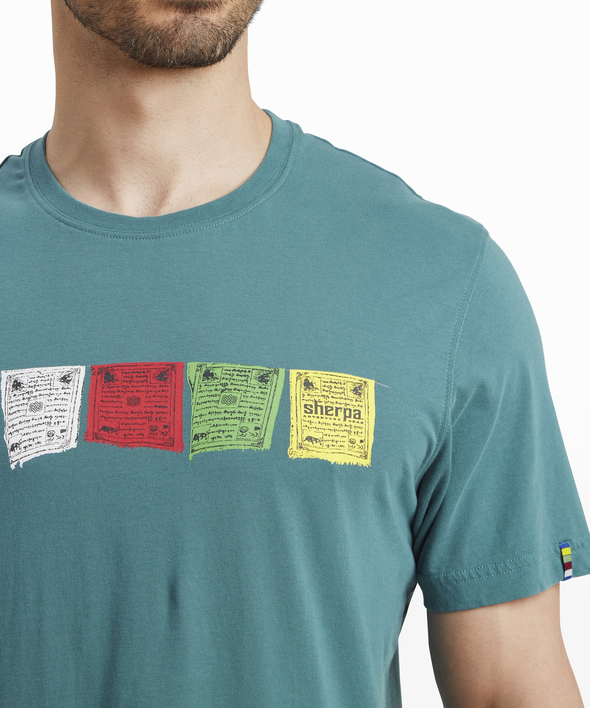 A close-up of the colorful Tibetan prayer flag design on the Sherpa Adventure Gear Tarcho T-Shirt in Blue. The flags are printed in blue, white, red, green, and yellow, each with intricate text and patterns, with the Sherpa logo visible on the yellow flag. A small multicolored detail is visible on the sleeve hem.