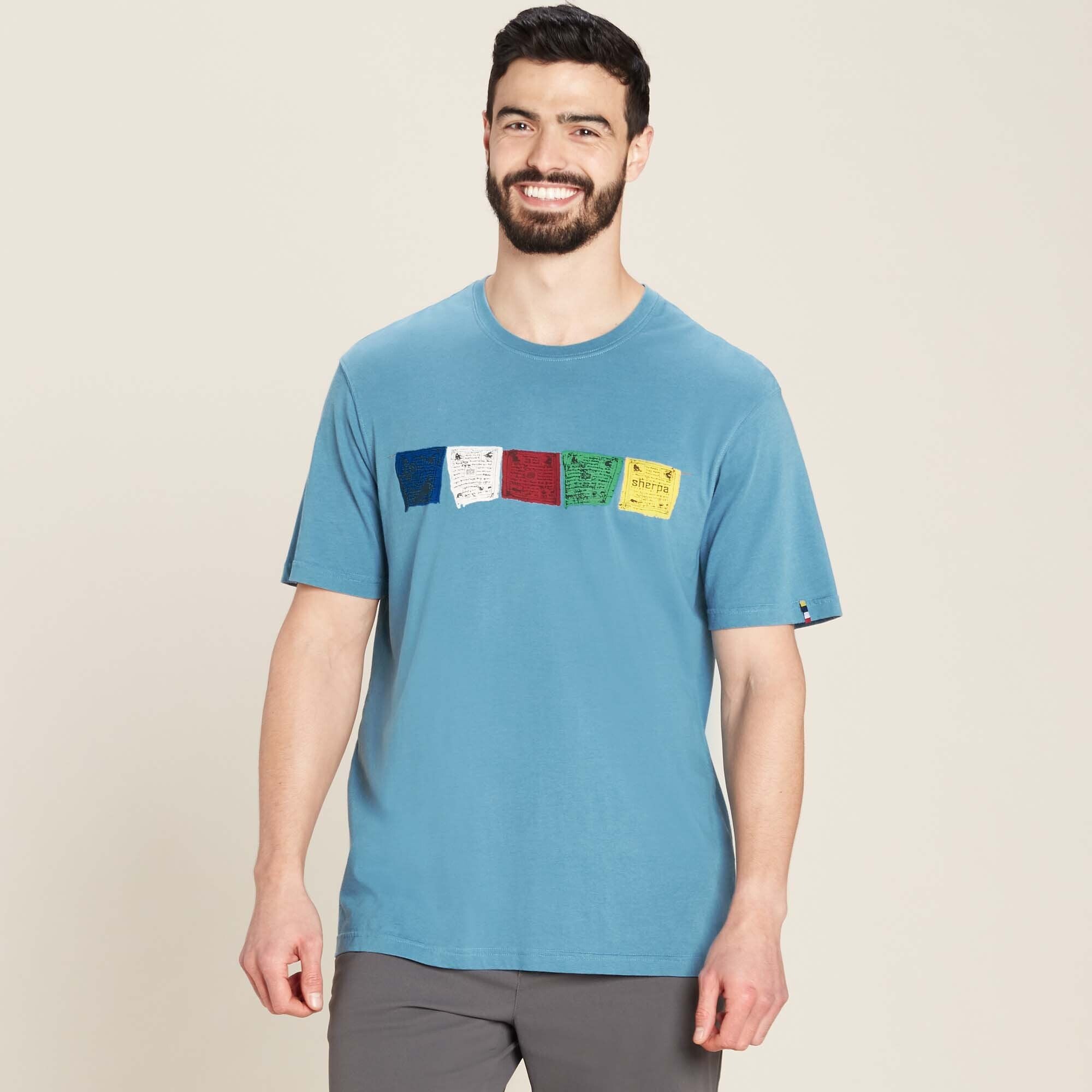 A male model with short dark hair, a beard, and an athletic build is wearing the Sherpa Adventure Gear Tarcho T-Shirt in Blue. The short-sleeve t-shirt features a horizontal row of printed Tibetan prayer flags across the chest in five colors: blue, white, red, green, and yellow. Each flag has traditional script and symbols printed on it. The model is smiling and standing with his hands at his sides. He pairs the shirt with grey pants. The shirt has a relaxed fit and a classic crew neck.