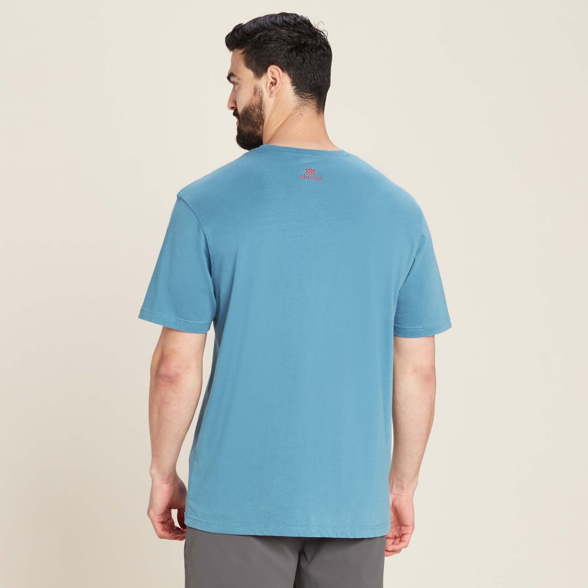 The same male model is shown from the back, wearing the Sherpa Adventure Gear Tarcho T-Shirt in Blue. The back of the t-shirt is solid blue with a small red Sherpa logo printed below the neckline. The fabric appears soft, with a natural drape. The sleeves fall just above the elbows, and the hem sits at the hips. The model’s posture is relaxed, with his hands slightly away from his body.