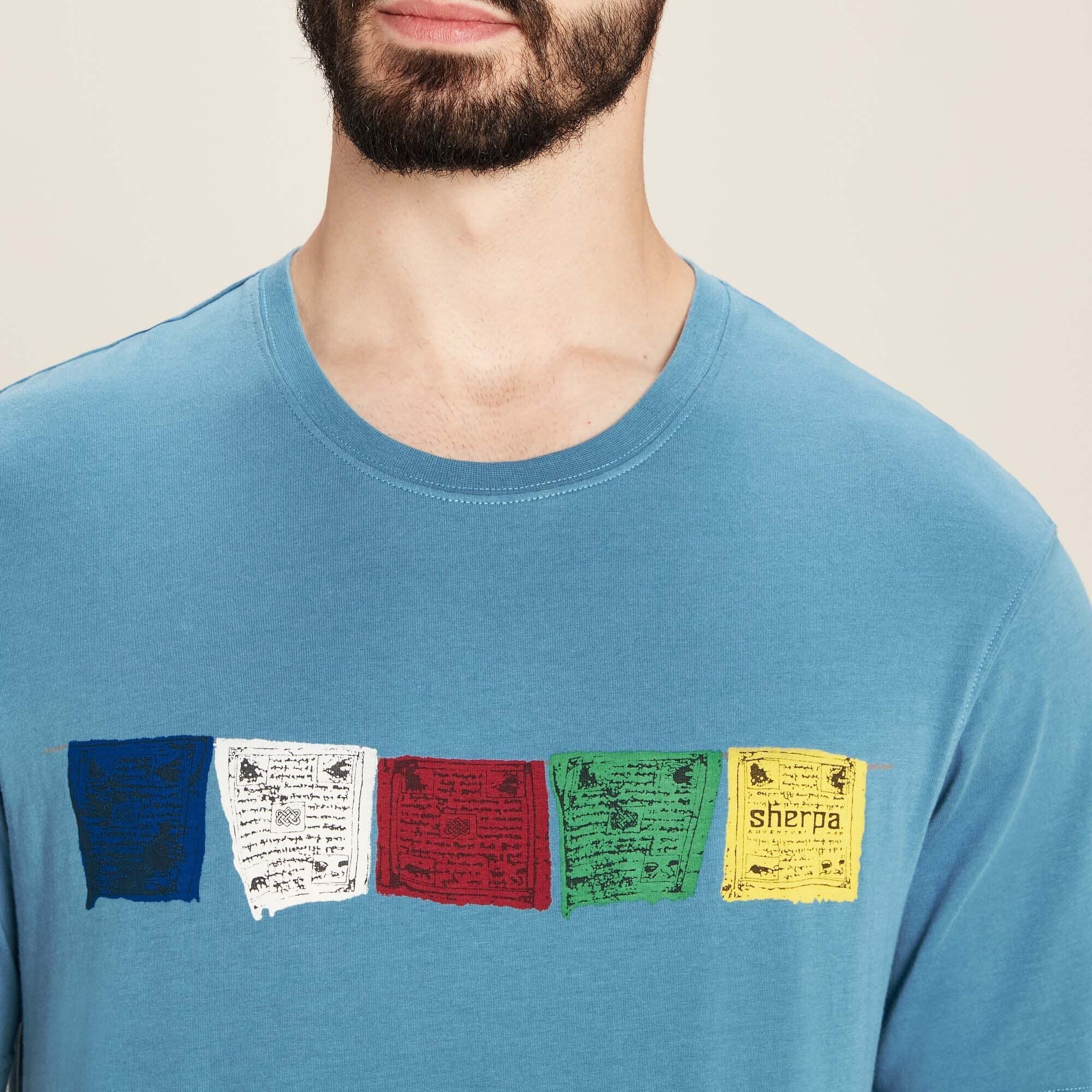 A close-up shot focuses on the chest area of the Sherpa Adventure Gear Tarcho T-Shirt in Blue, highlighting the colorful prayer flag design. Each flag displays intricate script and symbolic artwork, with the final yellow flag including the “Sherpa” brand name. The texture of the soft cotton fabric is visible, and the stitching around the neckline is clean and even. The prayer flag design adds a cultural and artistic touch to the simple t-shirt.