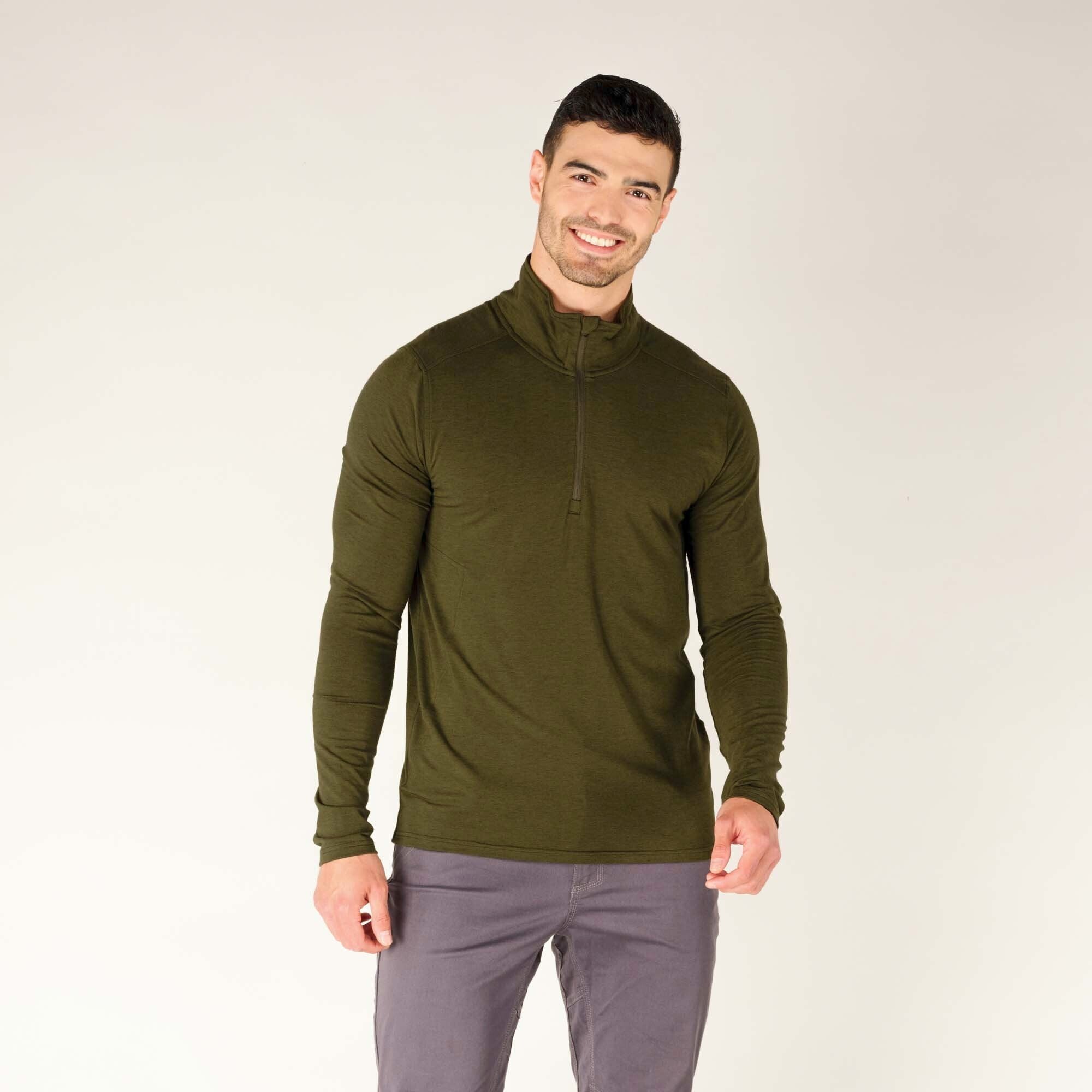 A male model with short dark hair and light skin is smiling while wearing a fitted Sherpa Adventure Gear Tendu Active Half Zip in Green with long sleeves. The pullover has a high collar with a front zipper extending down to the chest. He is also wearing gray pants and is posing against a plain beige background with a relaxed posture, with his hands slightly curled.