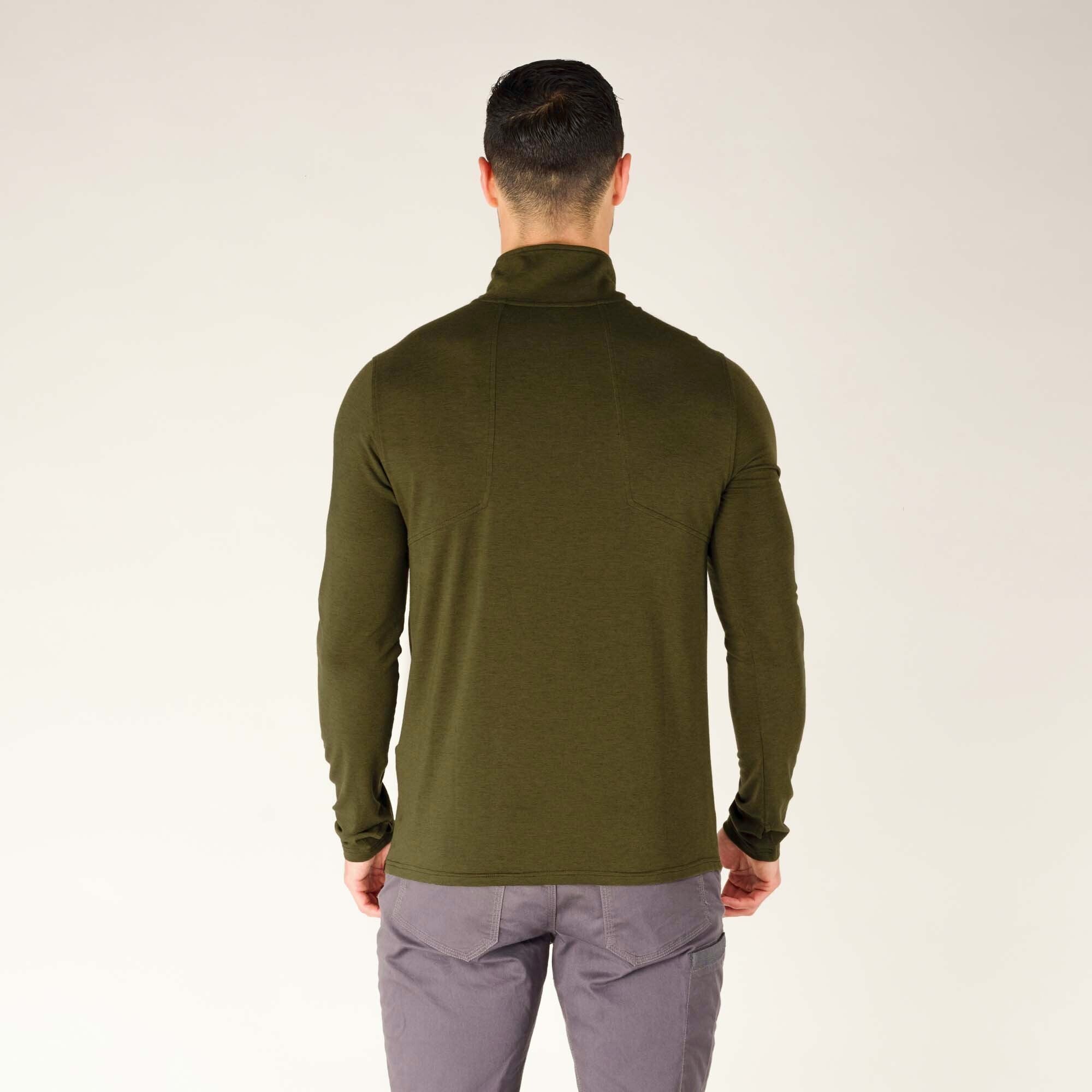 A full-body shot of the model from a back angle, showing the length of the Sherpa Adventure Gear Tendu Active Half Zip in Green extending just past his waist. His gray pants complement the olive-green color of the top. He is wearing brown hiking-style shoes with a rugged sole.