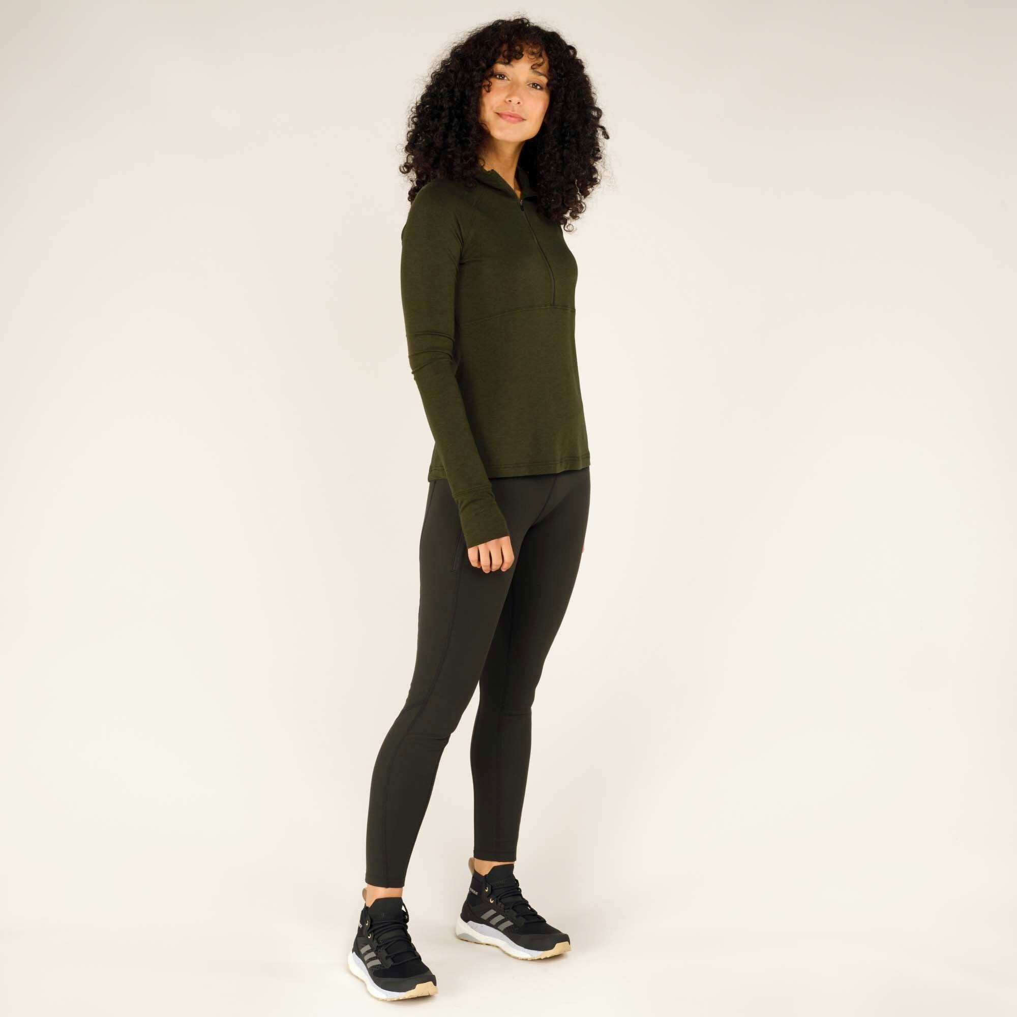 The same woman is shown in a full-body shot wearing the Sherpa Adventure Gear Tendu Active Half Zip in Green, paired with black fitted leggings and black Adidas trainers with a white sole. She is in a relaxed stance with one foot slightly in front of the other and looking slightly off to the side.