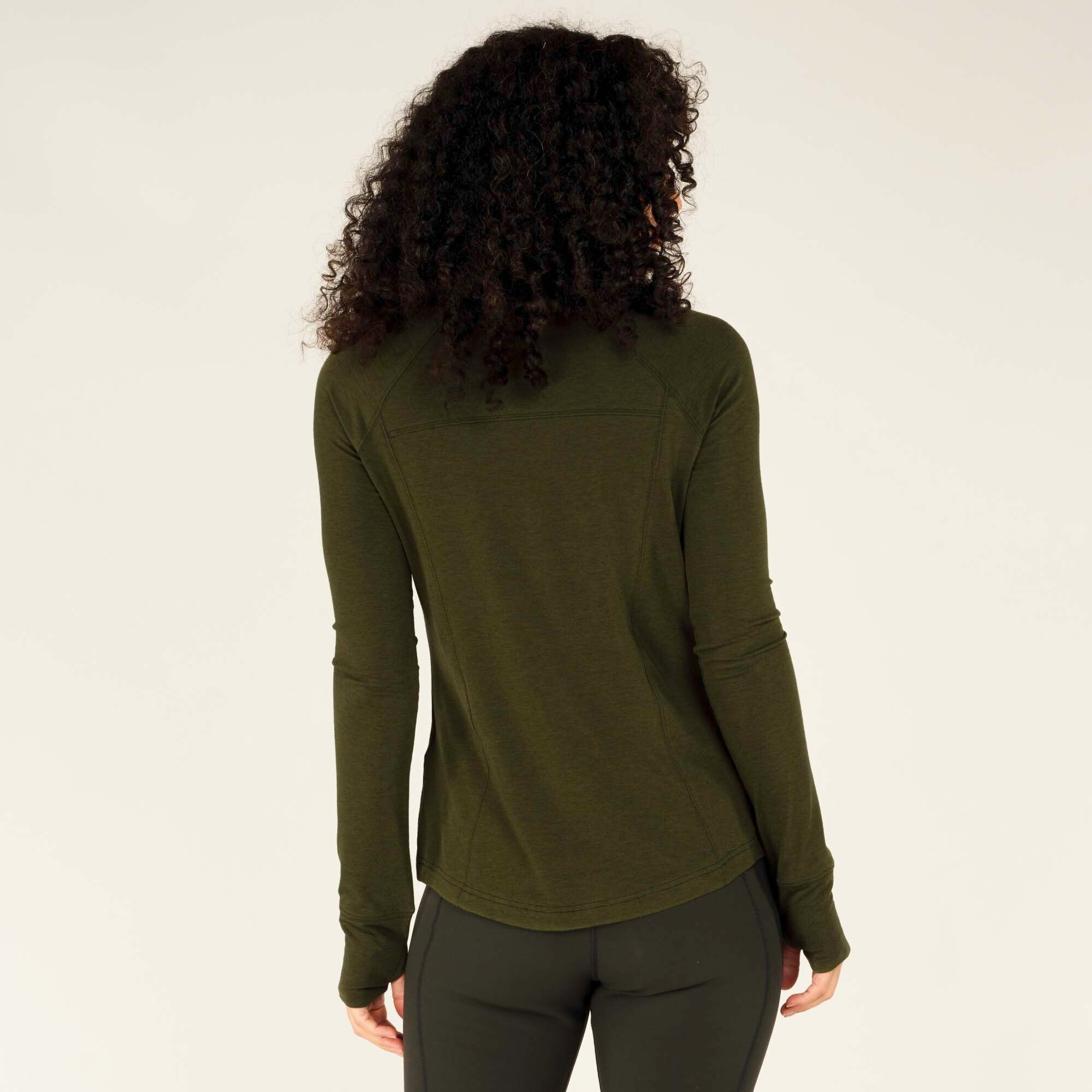 A back view of the woman showcasing the athletic top’s design. The Sherpa Adventure Gear Tendu Active Half Zip in Green has a curved hem and subtle stitching details along the upper back. The fitted sleeves include thumbholes, and she stands with her arms relaxed by her sides.