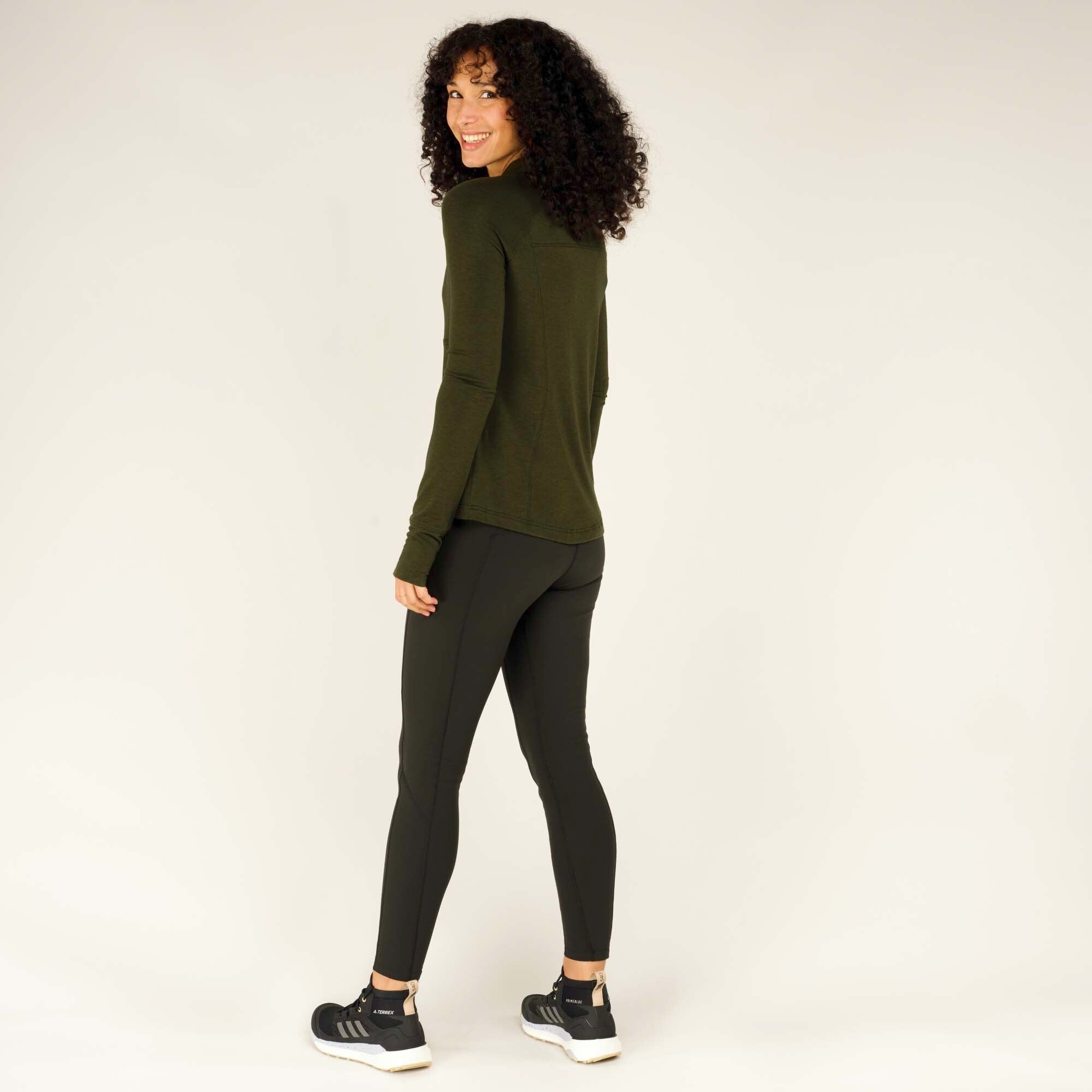 A slightly angled back view of the woman turning her head over her shoulder with a soft smile. The Sherpa Adventure Gear Tendu Active Half Zip in Green is shown from behind with the same fitted black leggings and black trainers.