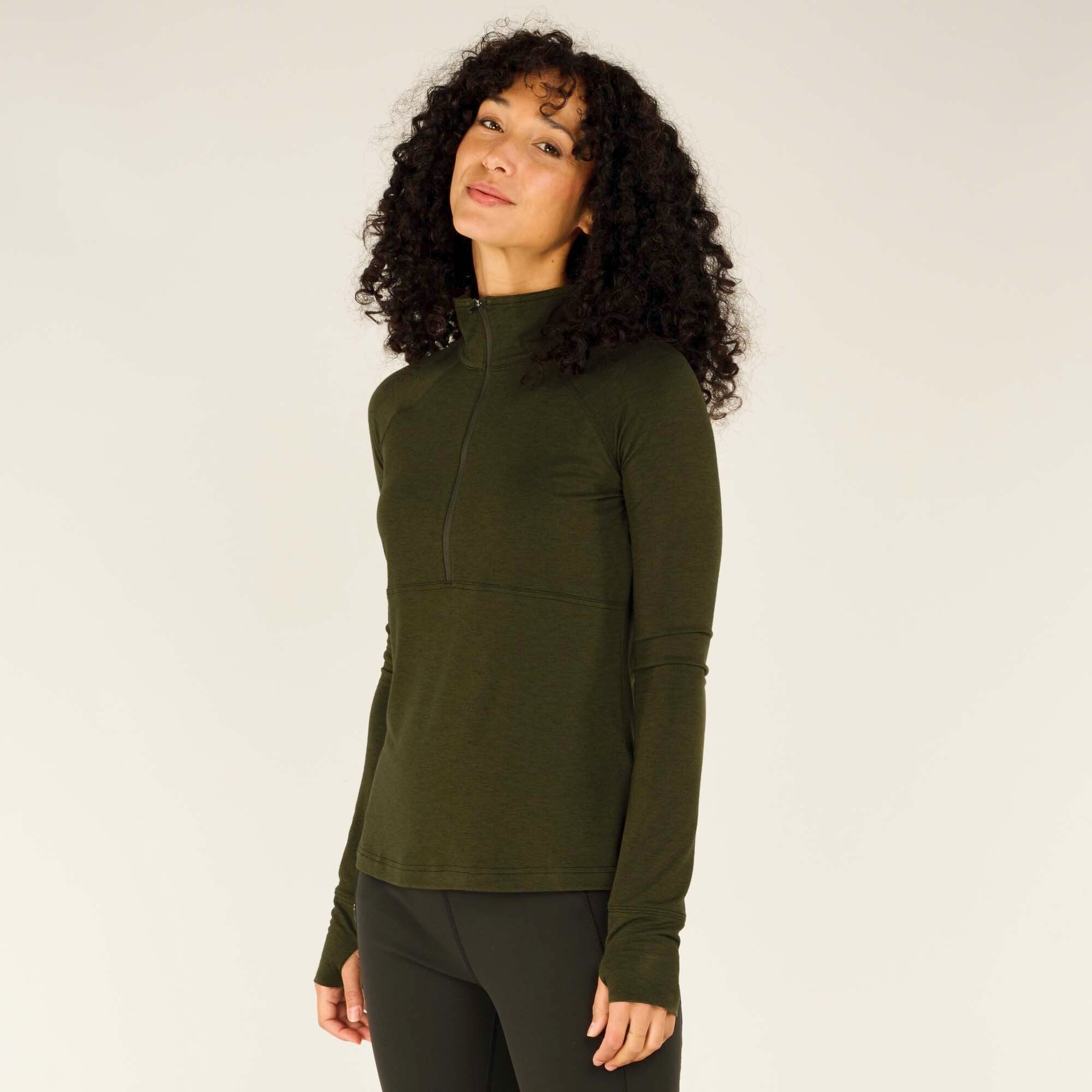 A closer front view of the woman wearing the Sherpa Adventure Gear Tendu Active Half Zip in Green with the collar zipped up to the top. Her hands are resting at her sides, and she has a confident yet relaxed expression. The long sleeves with thumbholes and the overall athletic fit of the top are clearly visible.