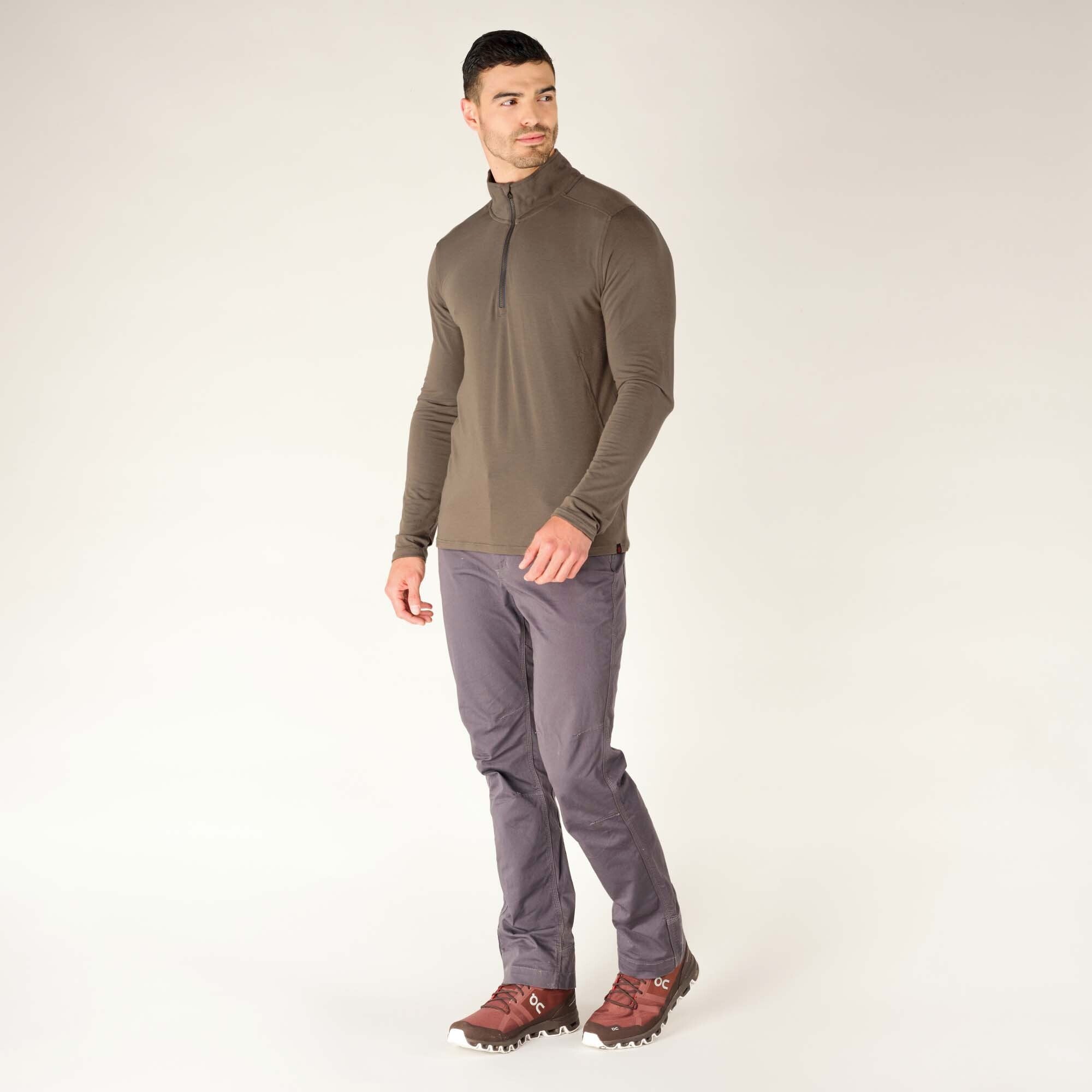 The model is now positioned with his side facing the camera, showing the side profile of the Sherpa Adventure Gear Tendu Active Half Zip in Grey. This angle highlights the smooth seamlines running down the sides and sleeves. He is wearing matching grey pants and brown hiking shoes, giving the outfit an outdoor adventure aesthetic. His hands are relaxed at his sides, and he is gazing off to the side.