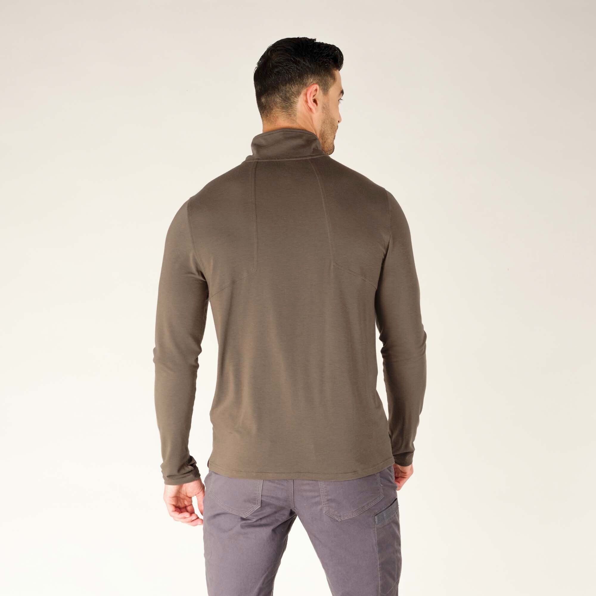 The model is standing with his back fully facing the camera, showcasing the back design of the Sherpa Adventure Gear Tendu Active Half Zip in Grey. The back features clean lines and minimal detailing, with subtle stitching near the shoulder blades for shaping. The high collar is folded up slightly, and the fabric drapes smoothly down the back. The model’s grey pants complete the cohesive, neutral-toned look.