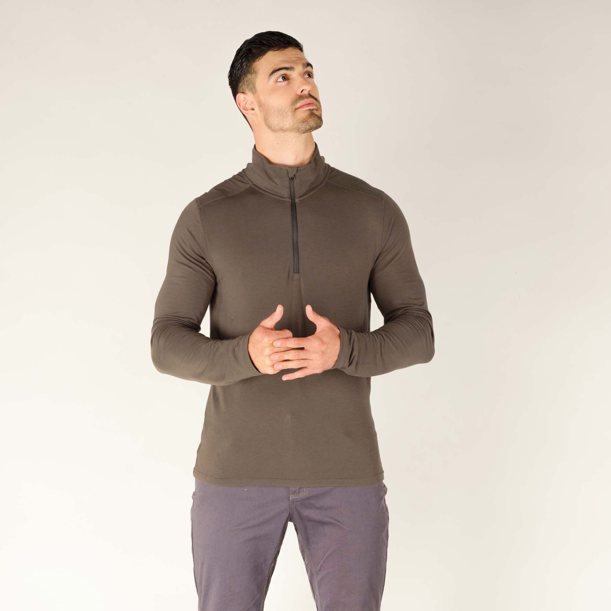 In this final image, the model is shown standing in a relaxed pose with his hands lightly clasped together at chest level, looking slightly upward with a thoughtful expression. This pose highlights the flexible fit of the Sherpa Adventure Gear Tendu Active Half Zip in Grey around the shoulders and arms, emphasizing its comfort and ease of movement. The grey half-zip’s sleek, minimal design is ideal for layering or standalone wear in outdoor or casual settings.