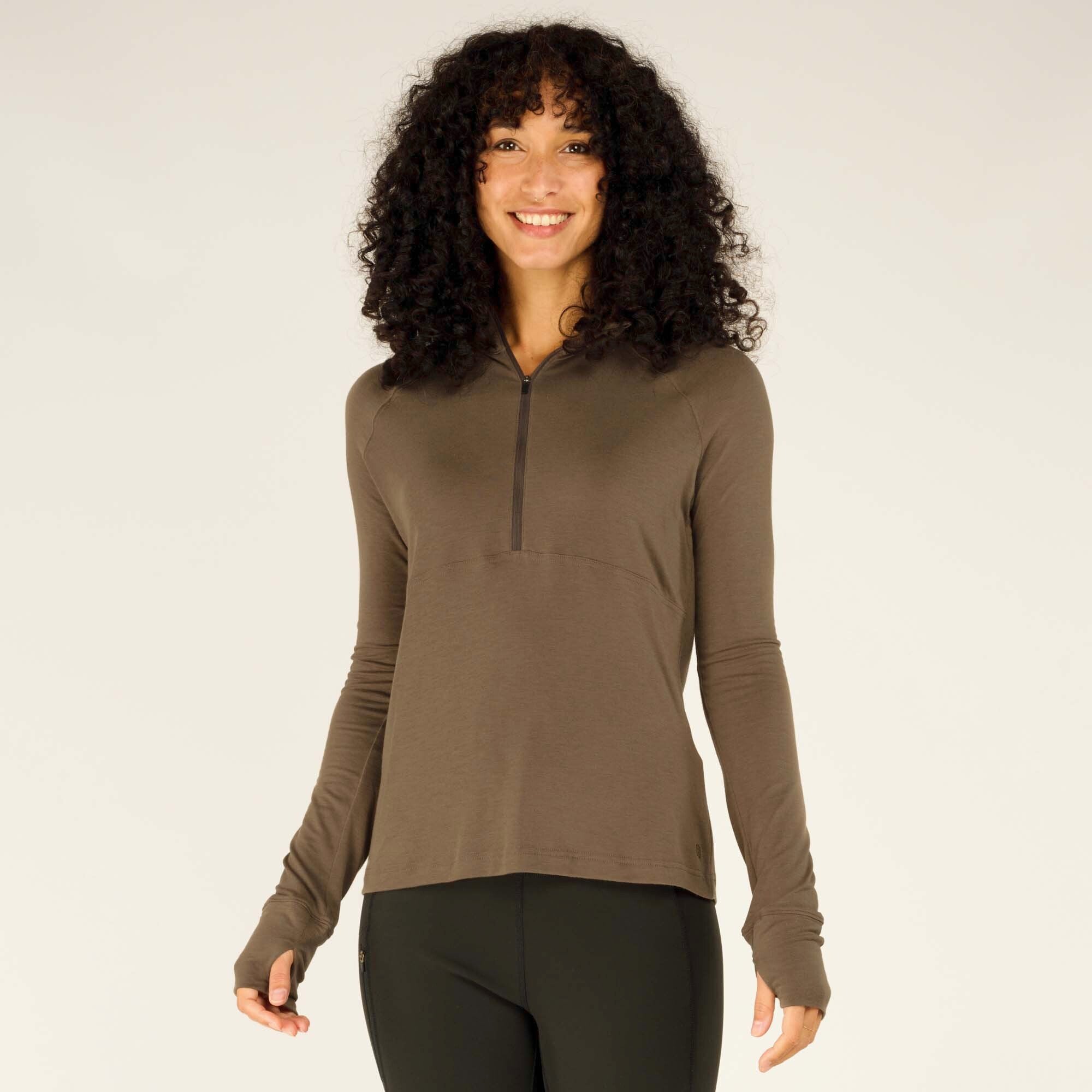 A woman with voluminous curly black hair smiles while wearing a fitted, long-sleeved, Sherpa Adventure Gear Tendu Active Half Zip in Grey. The lightweight fabric drapes comfortably, featuring a sleek quarter-zip at the collar. She pairs the top with black leggings that have a zippered pocket on the side.