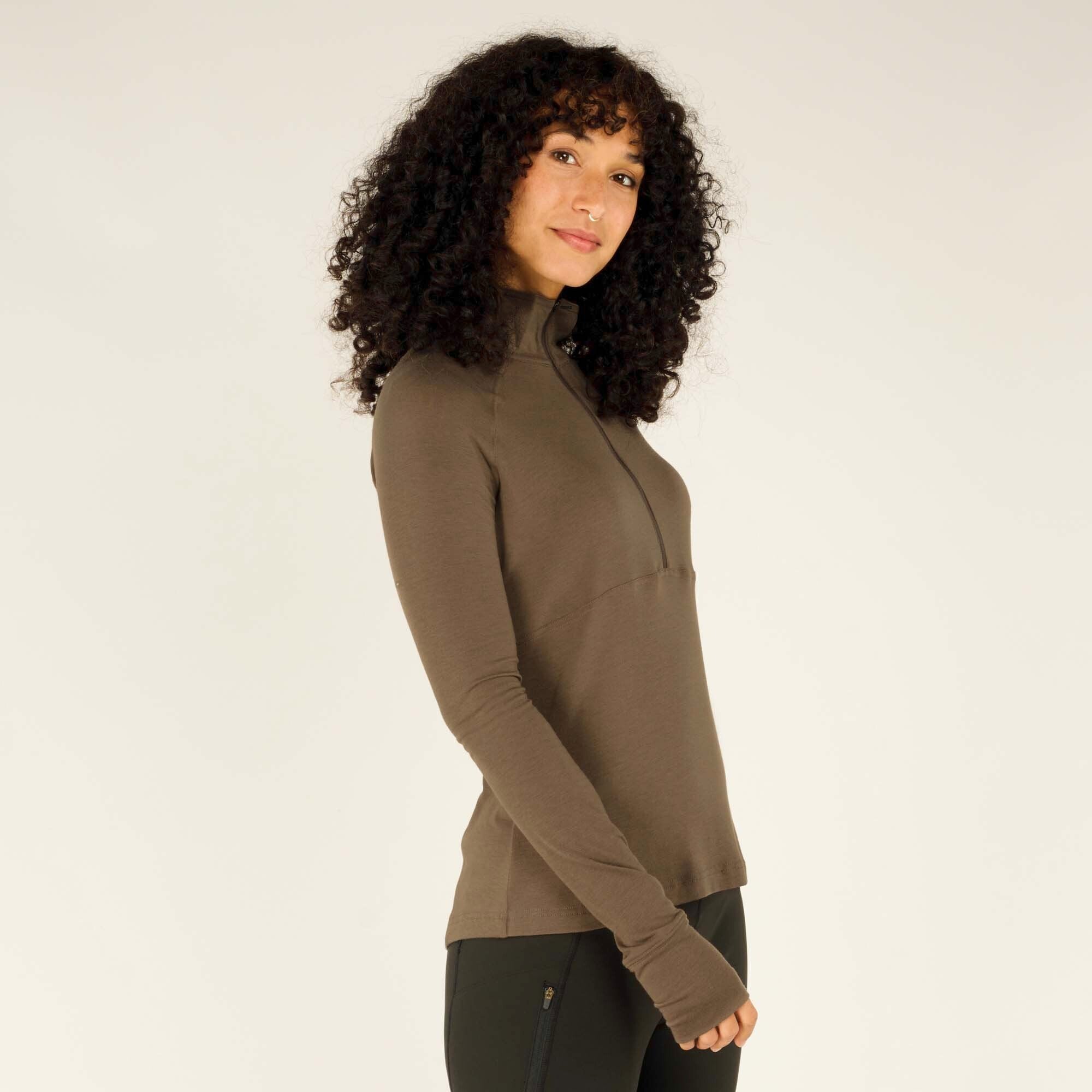 A closer shot of the woman, emphasizing the sleek fit of the Sherpa Adventure Gear Tendu Active Half Zip in Grey. The zipper is fully closed, showing how the collar sits against the neck. Her curly hair frames her face as she slightly tilts her head.