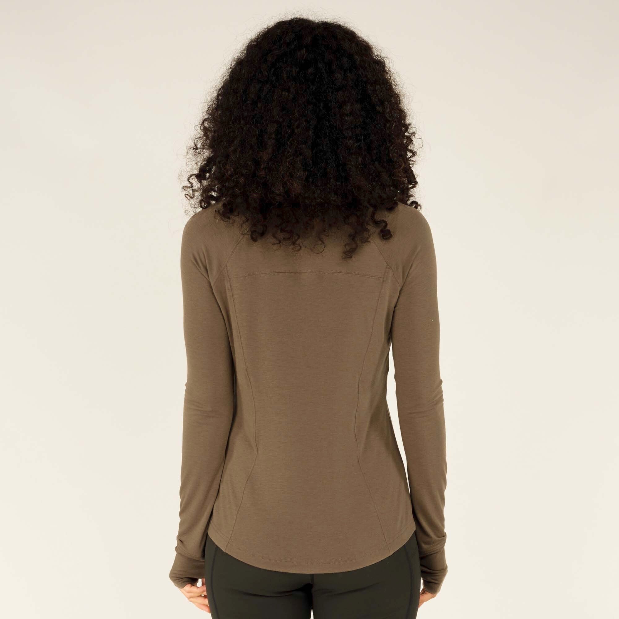 The woman is turned around, providing a clear view of the back of the Sherpa Adventure Gear Tendu Active Half Zip in Grey. The seams and structure of the top are visible, along with the slight curve of the hemline. The fabric appears smooth and stretchy, contouring to the body.