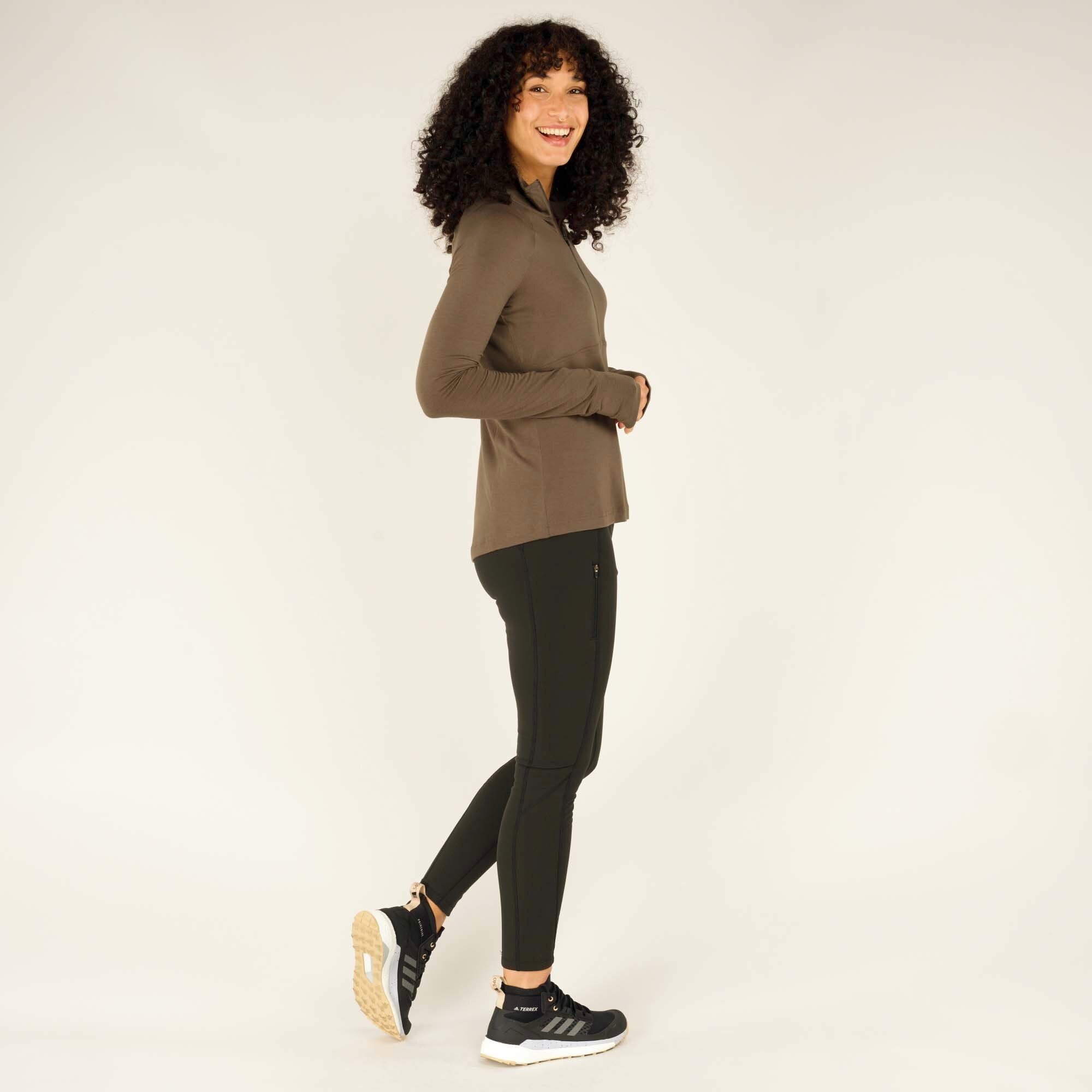 A dynamic shot capturing the woman mid-step. She looks over her shoulder with a bright smile, her movement showing the Sherpa Adventure Gear Tendu Active Half Zip in Grey’s flexibility. She wears black leggings and sporty sneakers, completing the activewear look.