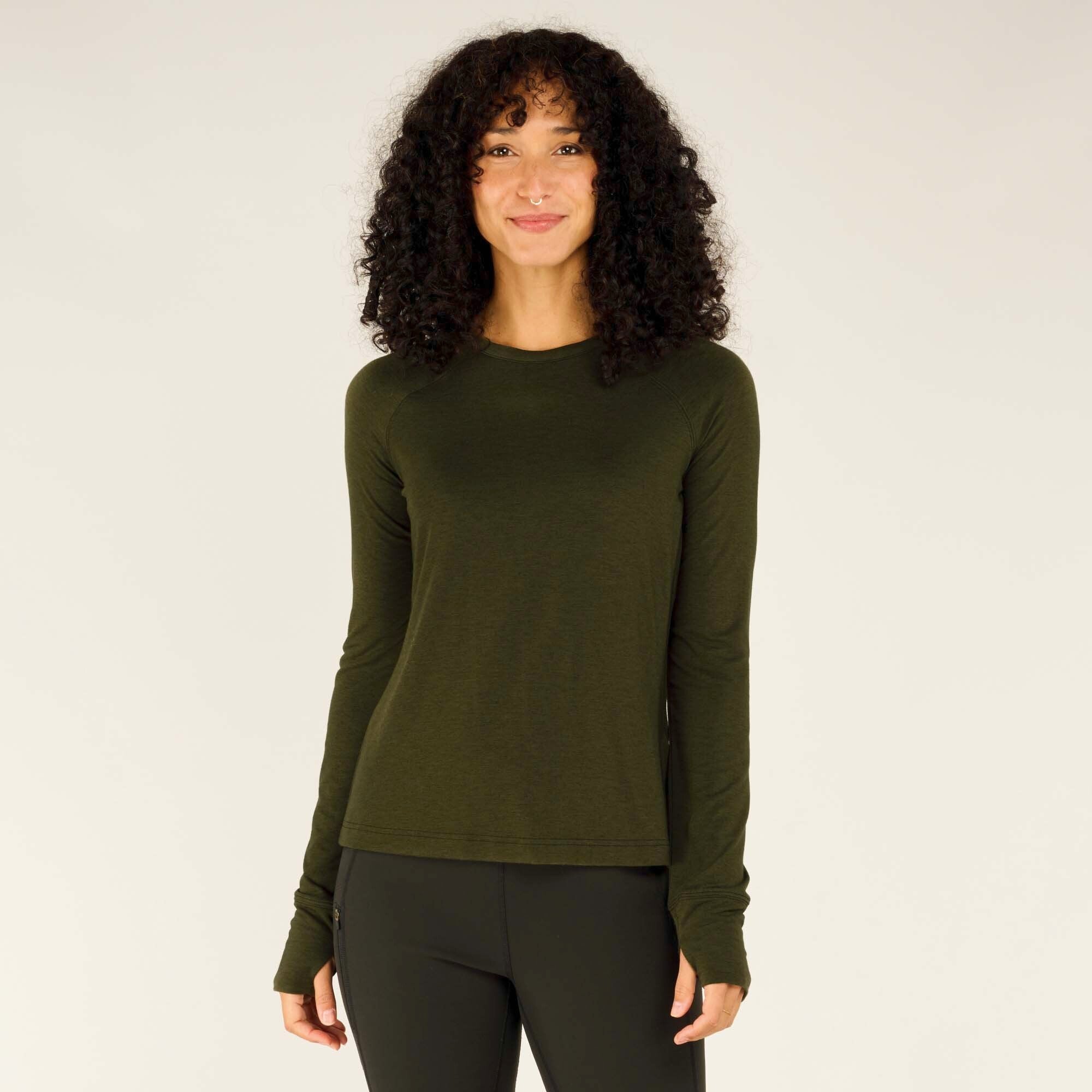 A young woman with curly black hair, a septum piercing, and a warm smile is modeling a Sherpa Adventure Gear Tendu Active Long Sleeve Crew in Green. The fitted athletic shirt has a subtle heathered texture, raglan sleeves, and thumbholes at the cuffs. She stands facing the camera with a relaxed posture, hands resting at her sides, and is wearing black leggings.