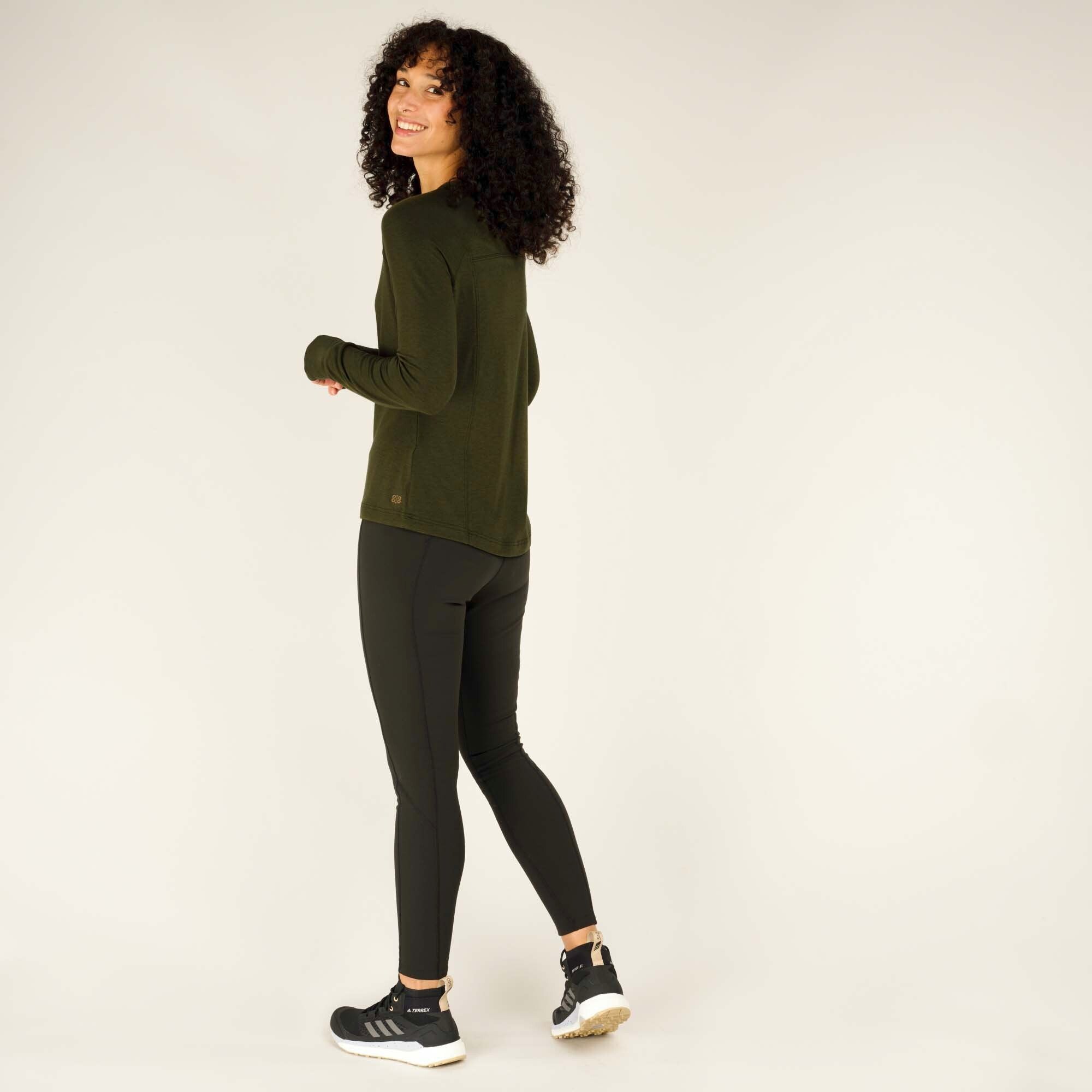 The same model is shown in a three-quarter rear view of the Sherpa Adventure Gear Tendu Active Long Sleeve Crew in Green, turning slightly to her right while smiling. The long-sleeve top has a slight curved hem, fitted sleeves, and reinforced stitching at the back yoke. She is paired with black leggings and black sneakers with white soles.