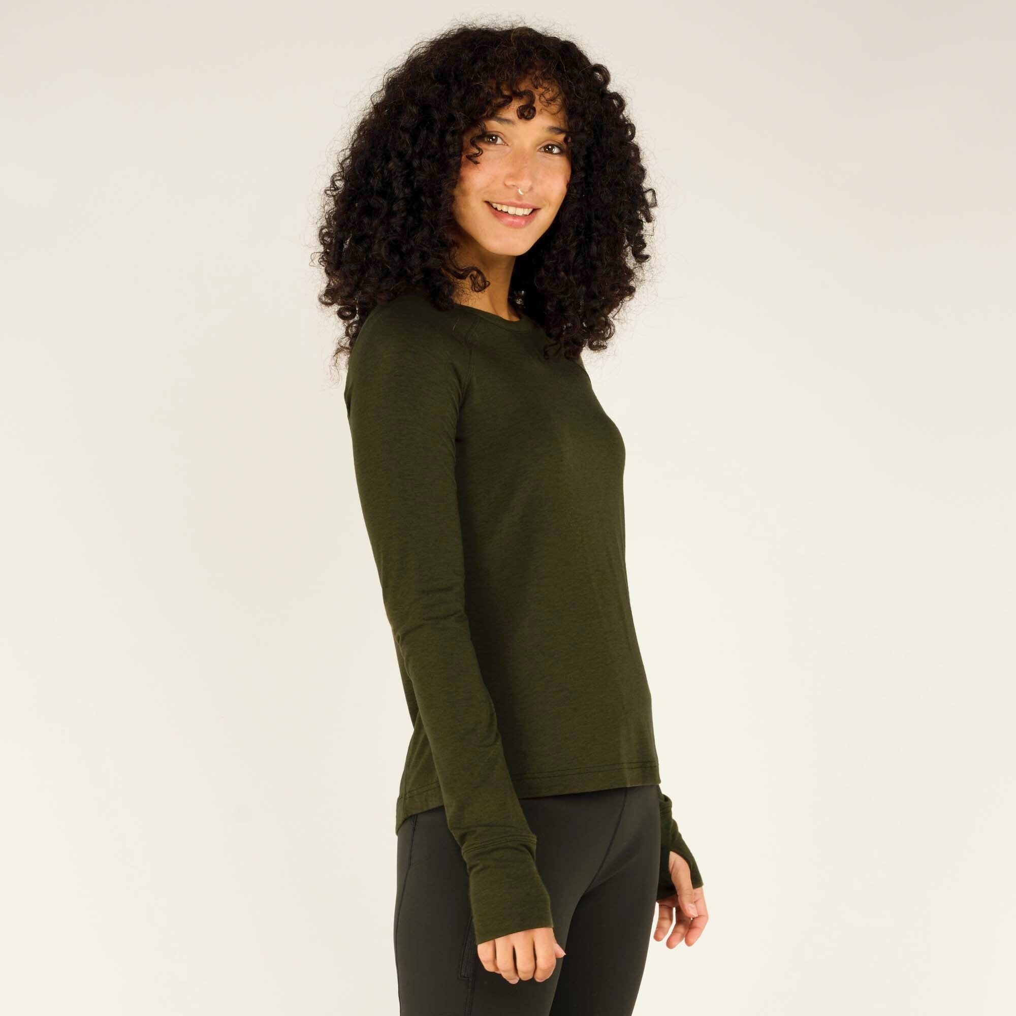 The model is captured in a profile shot, looking over her shoulder with a soft smile. The close-fitting fabric of the Sherpa Adventure Gear Tendu Active Long Sleeve Crew in Green follows the contours of her body, and the thumbholes at the sleeves are visible. The leggings have a smooth, high-waisted fit.