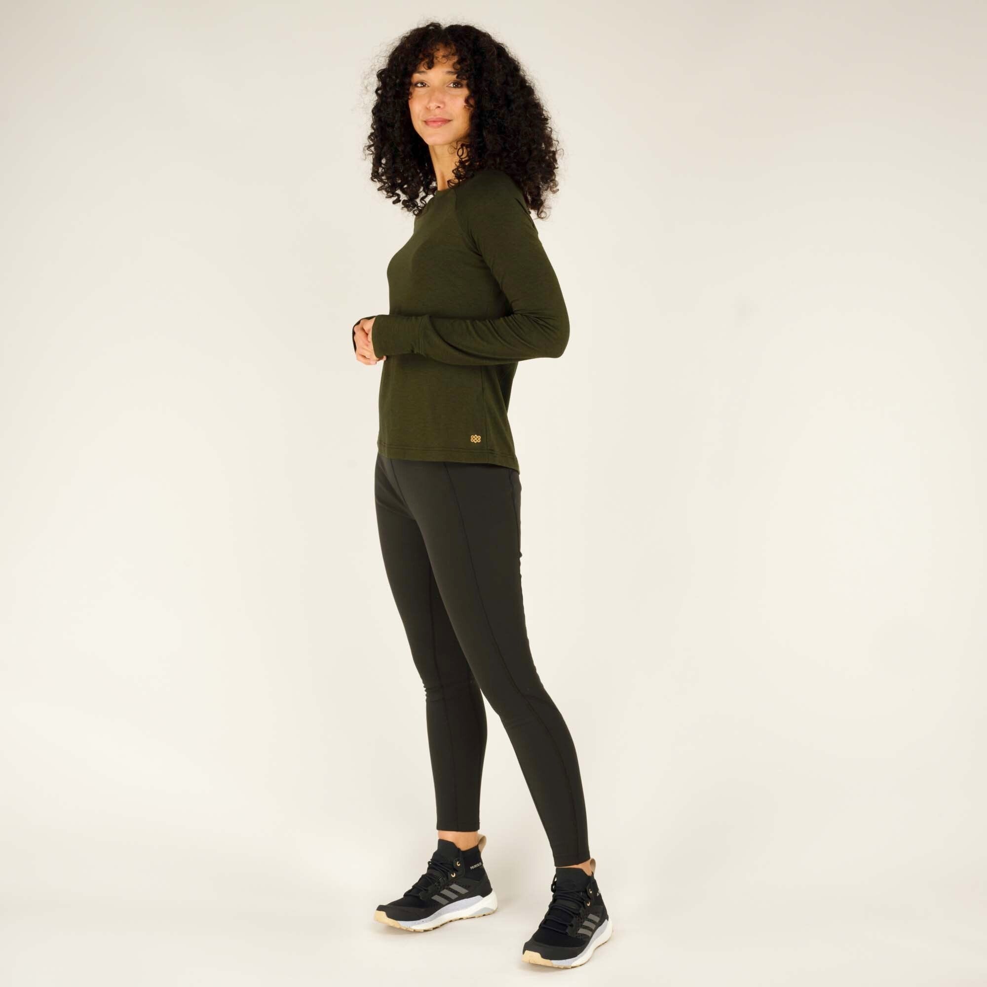 The model stands with her left leg slightly bent and her hands resting at her sides. She is wearing the Sherpa Adventure Gear Tendu Active Long Sleeve Crew in Green with black leggings and black sneakers with white soles. The fitted design of the shirt complements her form, and the lightweight material appears breathable.