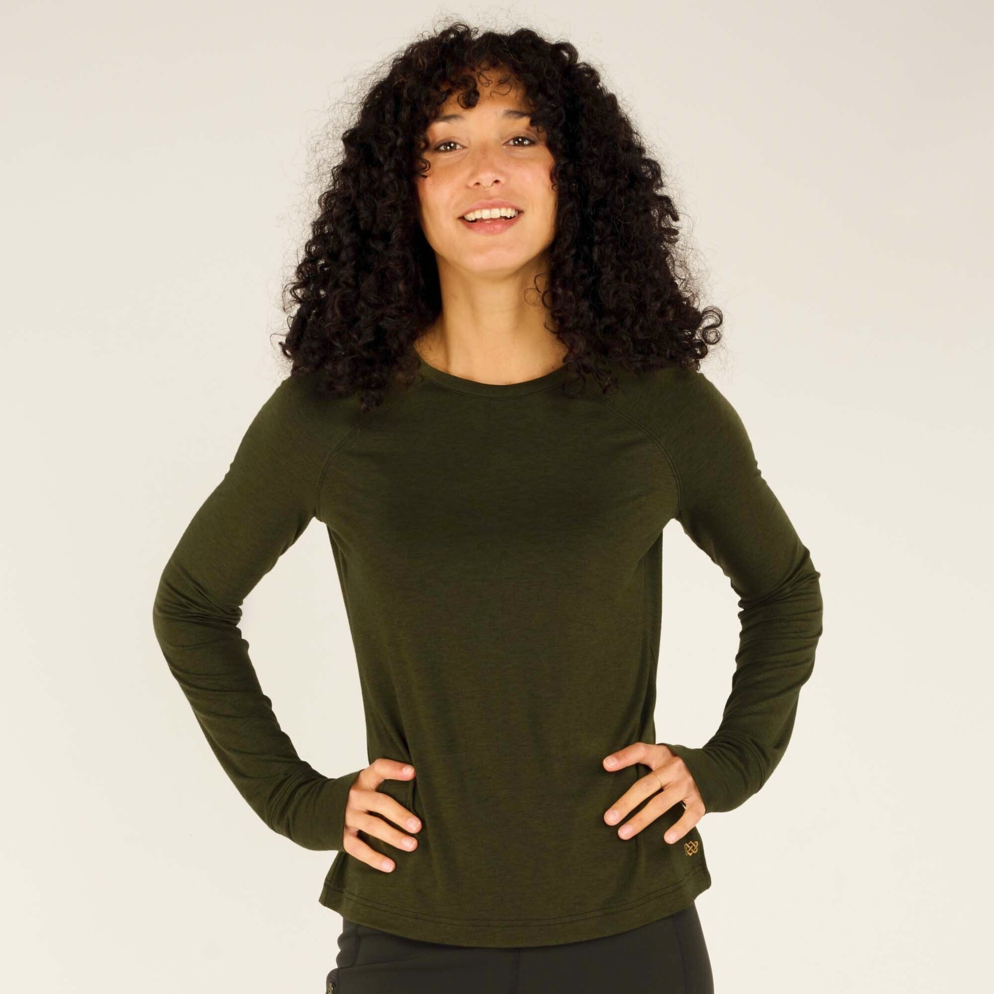 The model stands with her hands on her hips, giving a bright and confident smile. The Sherpa Adventure Gear Tendu Active Long Sleeve Crew in Green is snug but flexible, designed for active movement. The fabric's subtle texture and the small logo near the hem add detail to the minimalist design.