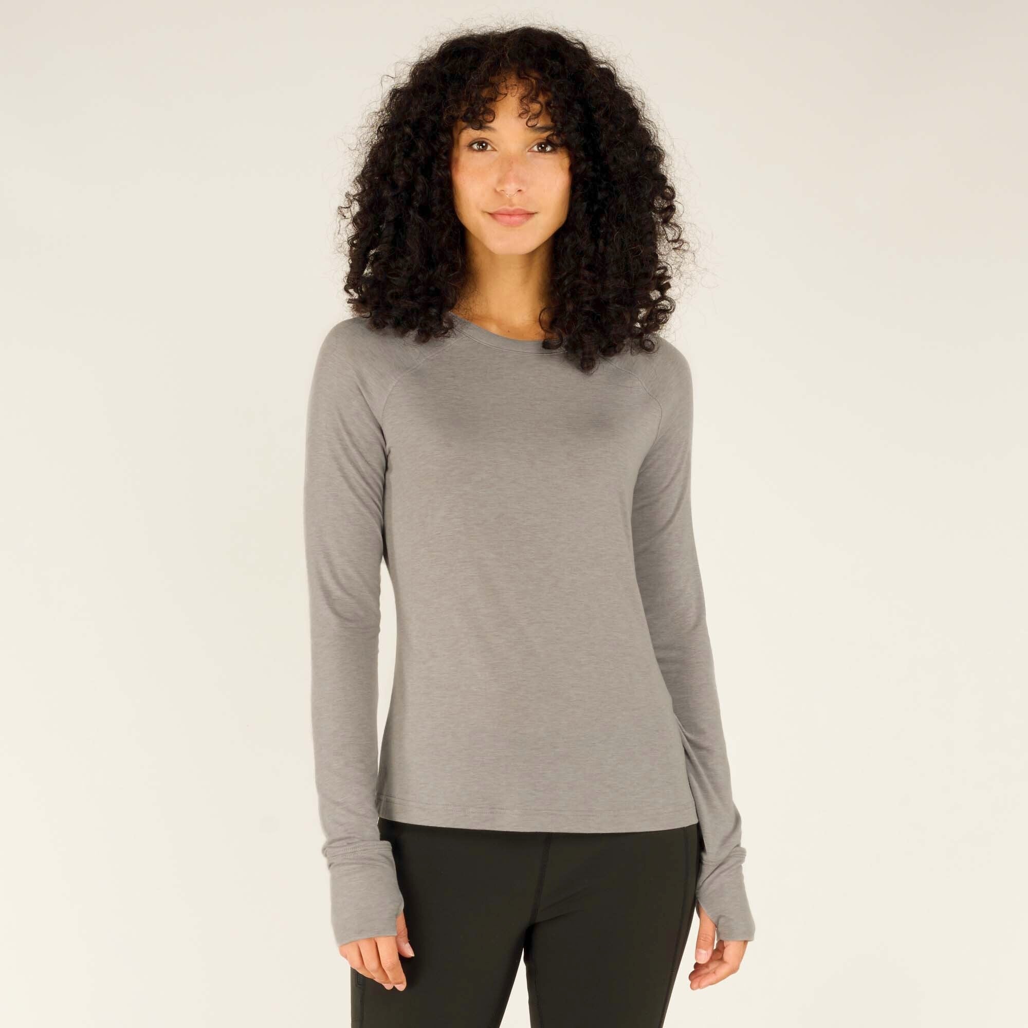 A female model with curly dark hair wears a fitted Sherpa Adventure Gear Tendu Active Long Sleeve Crew in Grey and black leggings, standing against a neutral background. She faces the camera directly with a soft smile, hands relaxed by her sides, showcasing the shirt's slim fit and thumbhole cuffs.