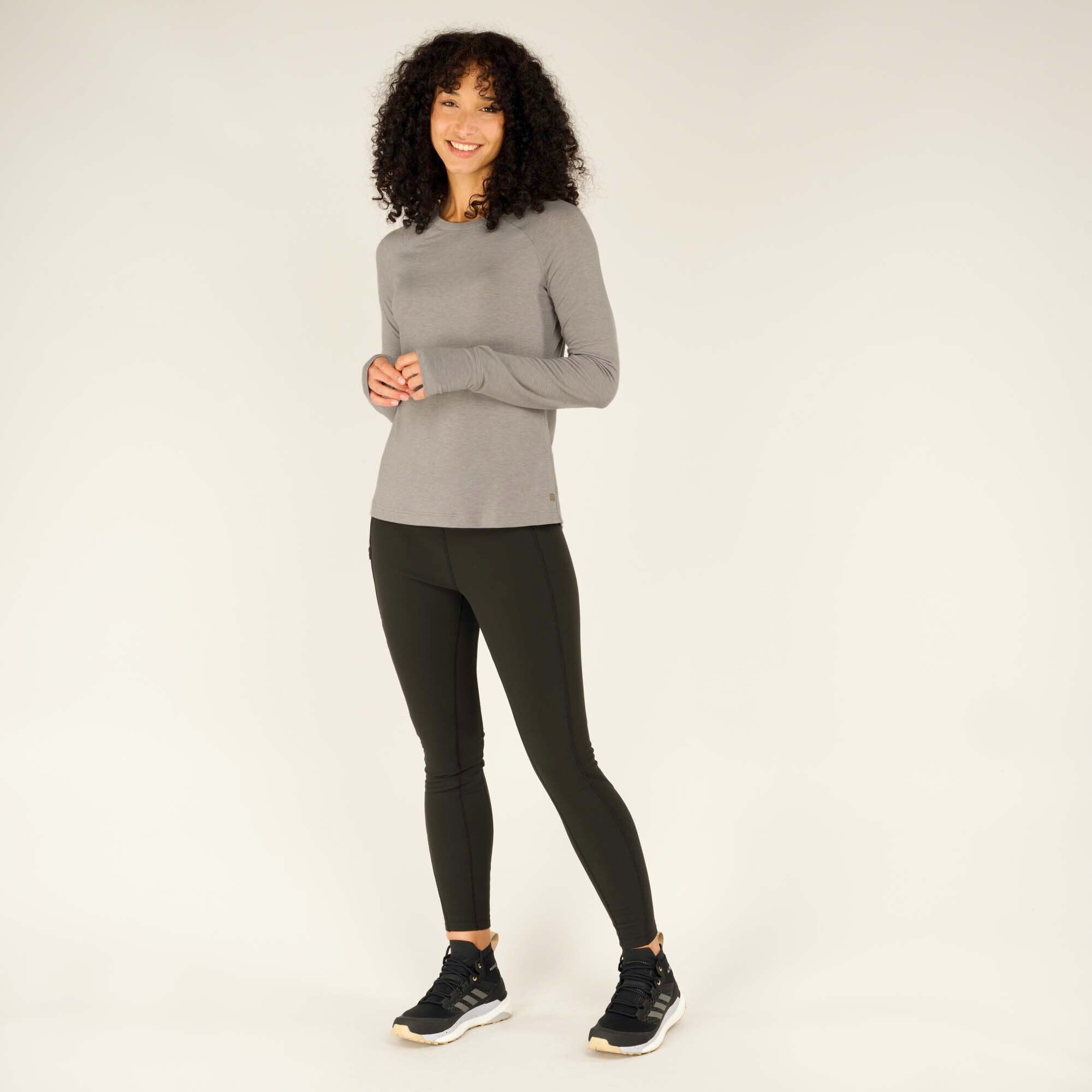 Full-body shot of the same model in the Sherpa Adventure Gear Tendu Active Long Sleeve Crew in Grey and black leggings. She stands in a relaxed pose with her hands gently clasped in front of her, smiling. The shirt’s slightly curved hem and streamlined fit are visible, along with her athletic black sneakers.