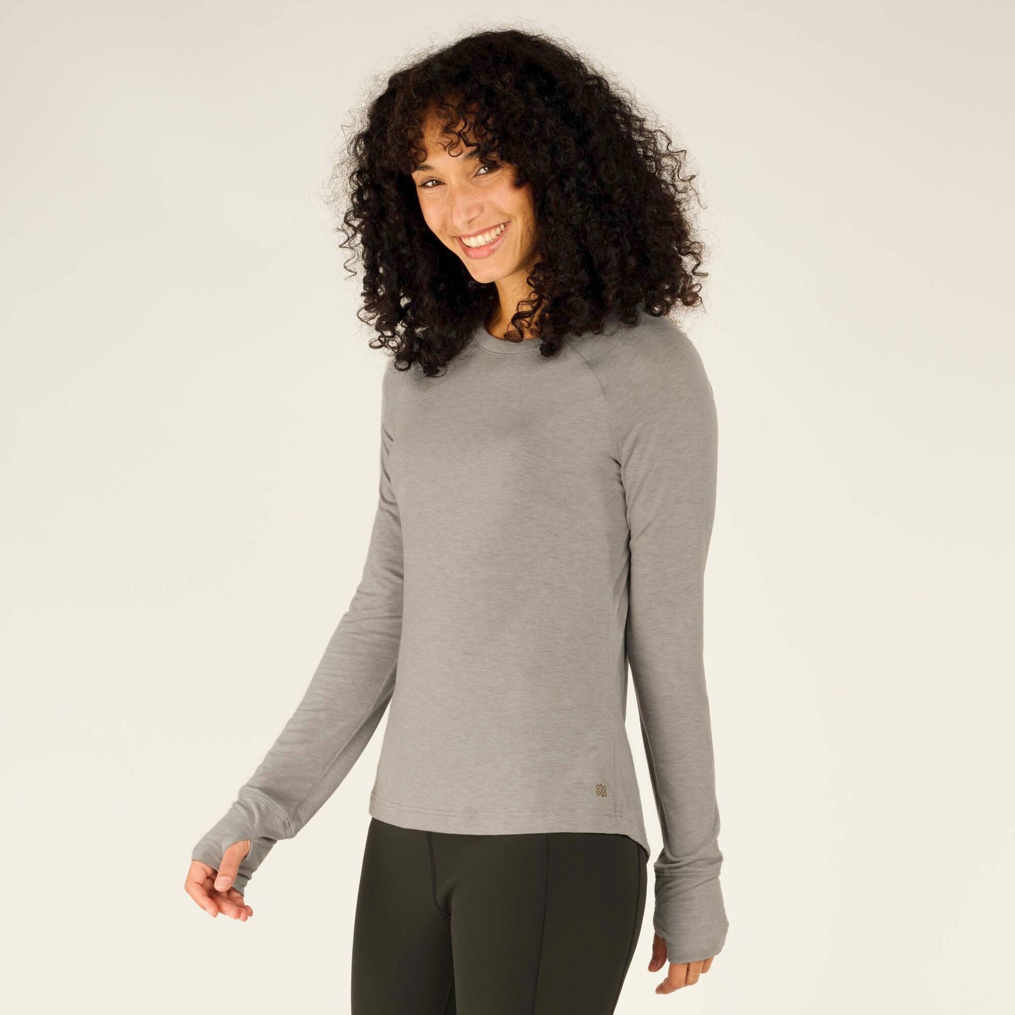 The model is shown from a three-quarter angle, turning slightly to her left, showing the side fit of the Sherpa Adventure Gear Tendu Active Long Sleeve Crew in Grey. She smiles brightly, and the close fit of the sleeves and body is apparent.