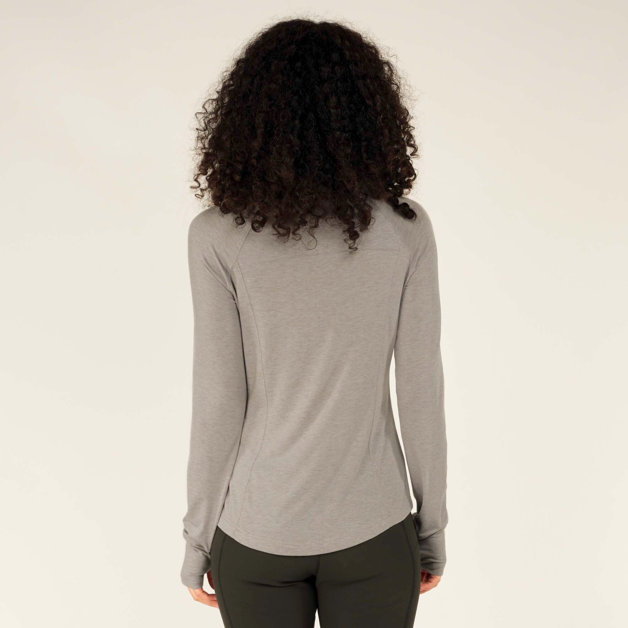 Back view of the model wearing the Sherpa Adventure Gear Tendu Active Long Sleeve Crew in Grey and black leggings. The shirt’s back seam details, fitted shape, and subtle curve at the hem are clearly visible. Her hair cascades down, partially covering the top.