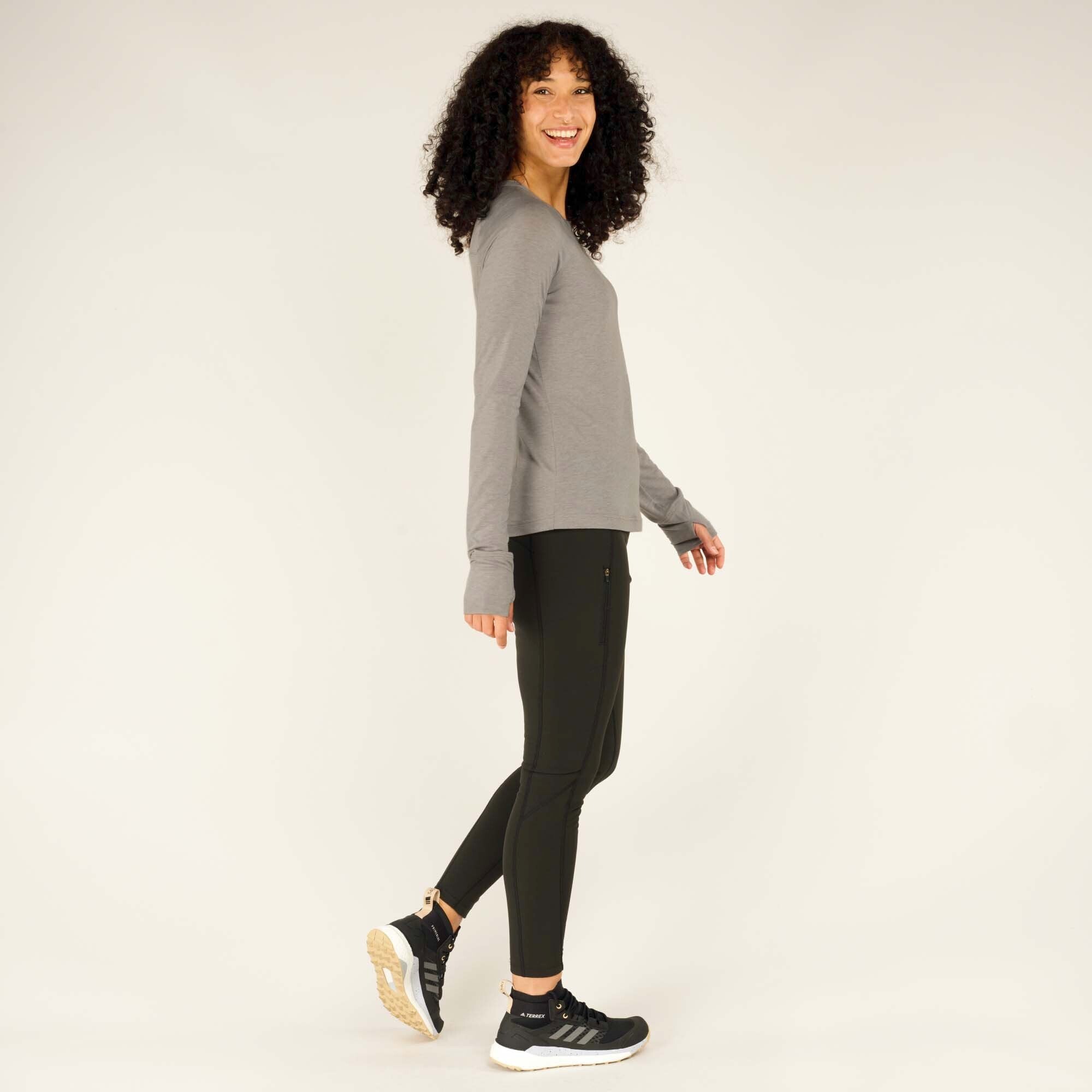 Side view of the model walking to her left while wearing the Sherpa Adventure Gear Tendu Active Long Sleeve Crew in Grey and black leggings. Her right foot is slightly lifted, showing the sole of her black sneakers with beige accents. The movement highlights the flexibility and comfort of the activewear ensemble.