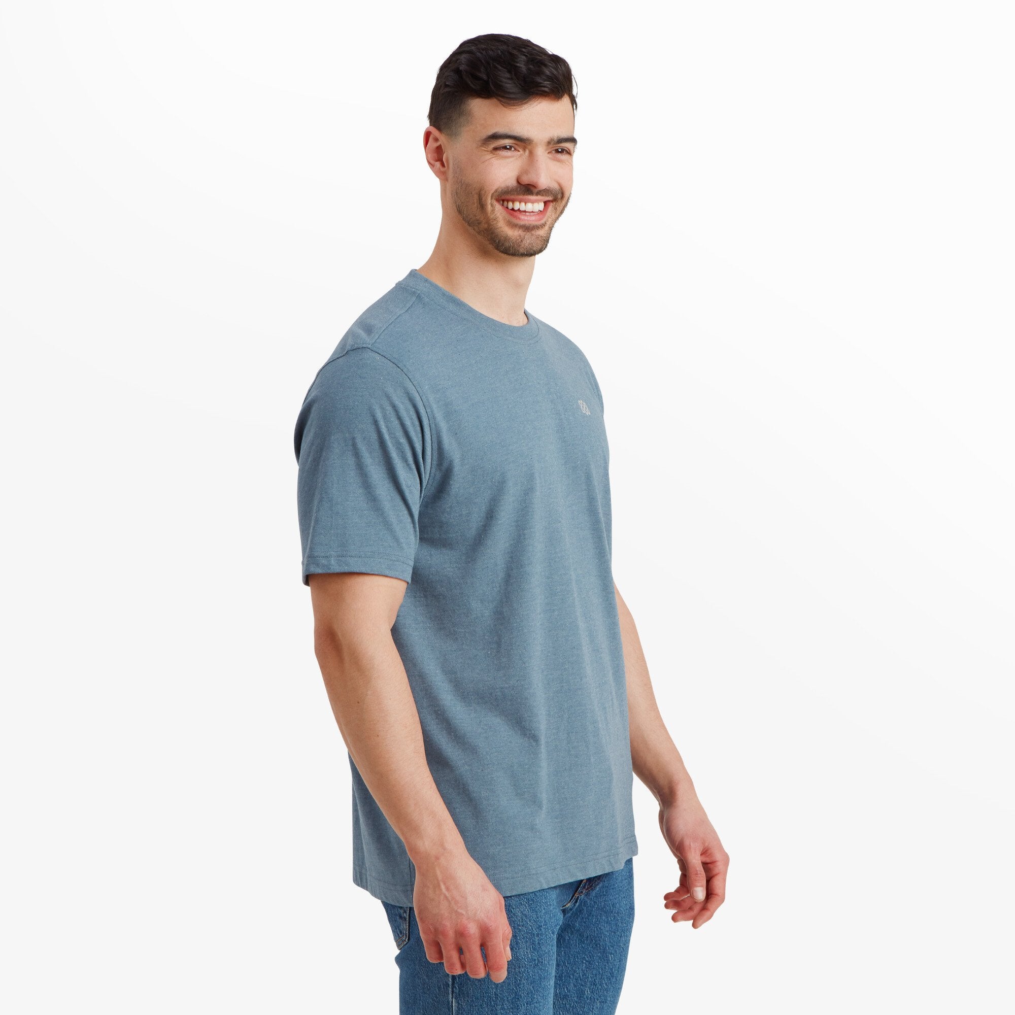 The same man as in the previous image, this time in a profile shot. He is slightly angled to the side, showcasing the relaxed fit of the Sherpa Adventure Gear Terrain T-Shirt in Blue. The Sherpa knot emblem is visible on the chest, with the hemline falling neatly over his jeans.