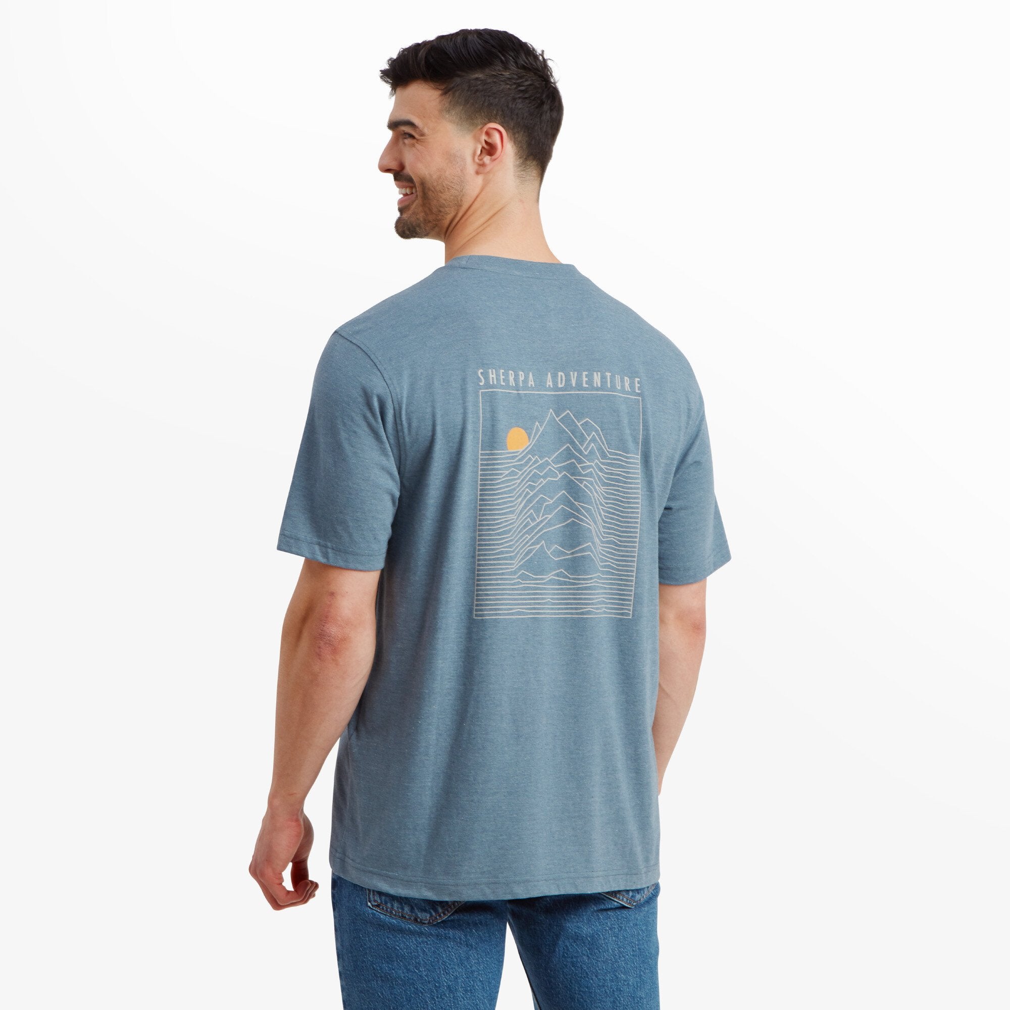 A back view of the man, highlighting the back design of the Sherpa Adventure Gear Terrain T-Shirt in Blue, which features a line-art depiction of mountains, a rising sun, and the text "Sherpa Adventure" in a clean font above the design. The design is printed in a light shade that complements the T-shirt color.