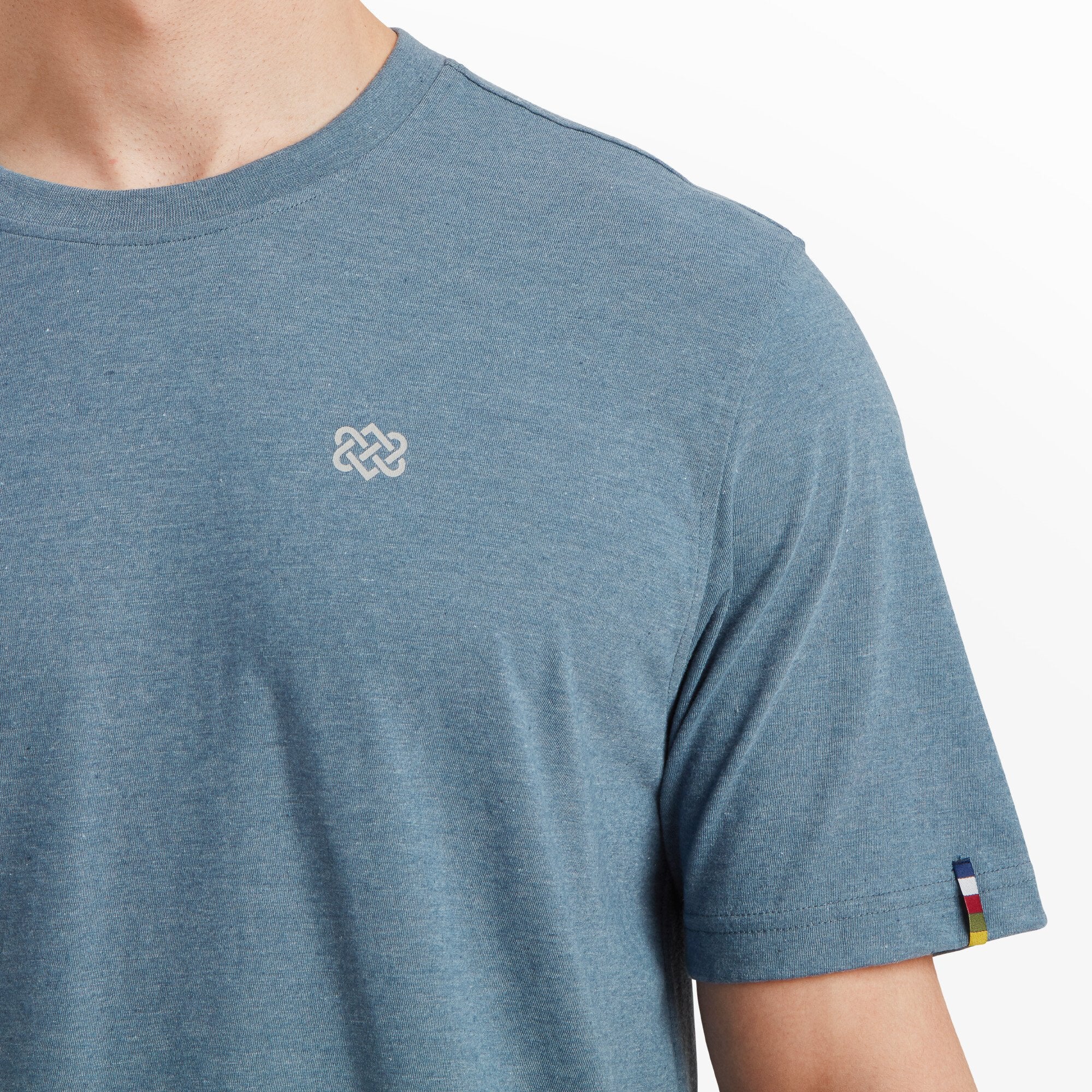 A close-up view of the front of the Sherpa Adventure Gear Terrain T-Shirt in Blue, focusing on the embroidered Sherpa knot emblem on the chest. The fabric texture is visible, with the soft blue tone standing out against the clean background.