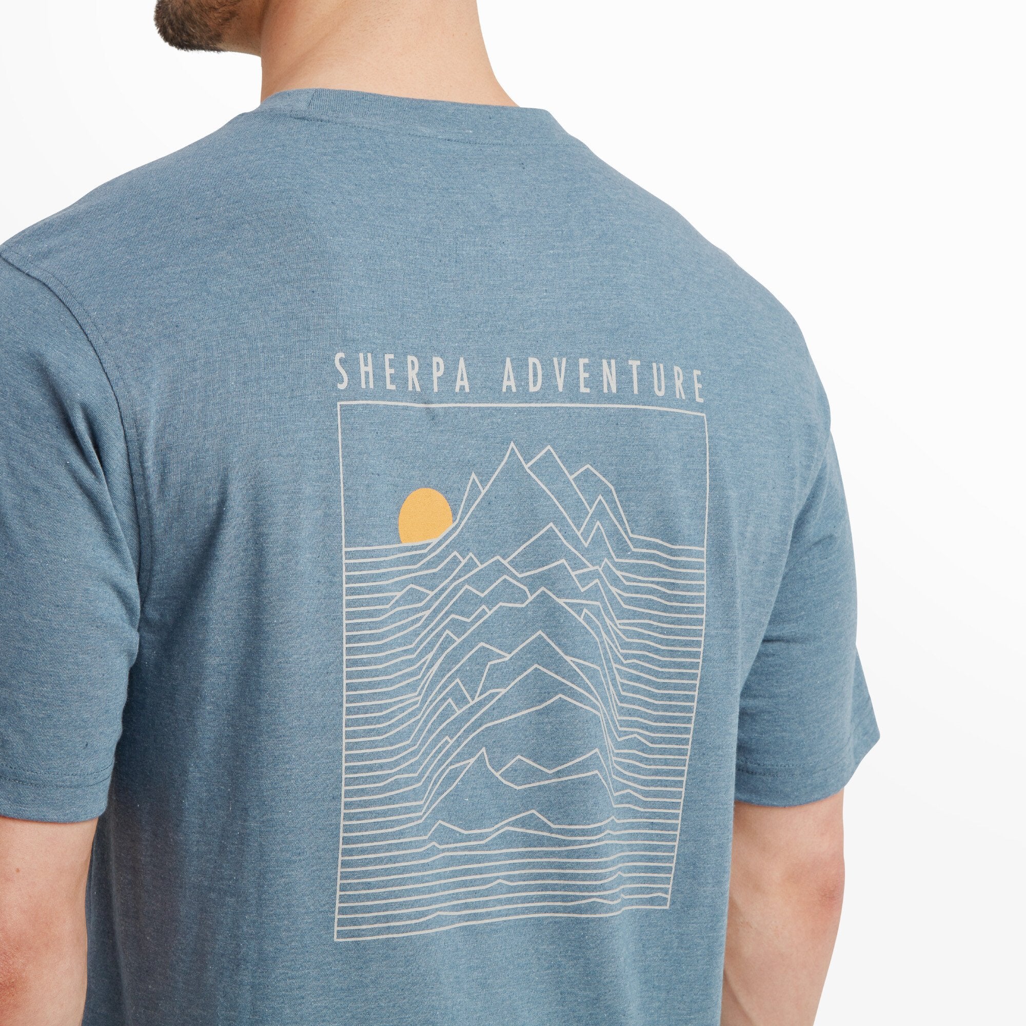 A close-up view of the back design of the Sherpa Adventure Gear Terrain T-Shirt in Blue, showing detailed line art of a mountain range and a rising sun within a rectangular frame. The text "Sherpa Adventure" is prominently displayed above the artwork. The texture of the fabric and the vibrant colors of the print are evident.