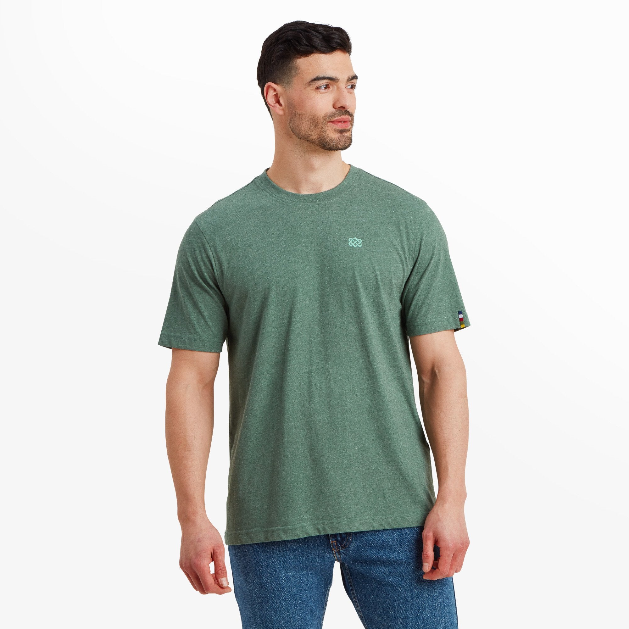 A man is wearing a Sherpa Adventure Gear Terrain T-Shirt in Green. The design is minimal, with a small geometric Sherpa logo embroidered on the chest. The t-shirt has a relaxed fit and pairs well with blue jeans.