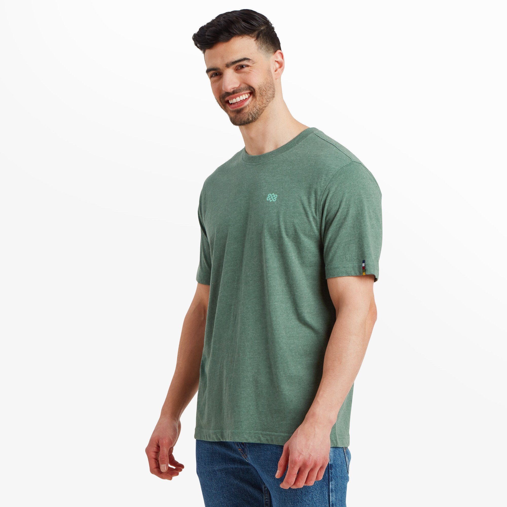 The model is turned slightly to the side, smiling while showcasing the Sherpa Adventure Gear Terrain T-Shirt in Green's comfortable and loose fit. The short sleeves are visible, highlighting the small colorful Sherpa flag detail on the left sleeve.