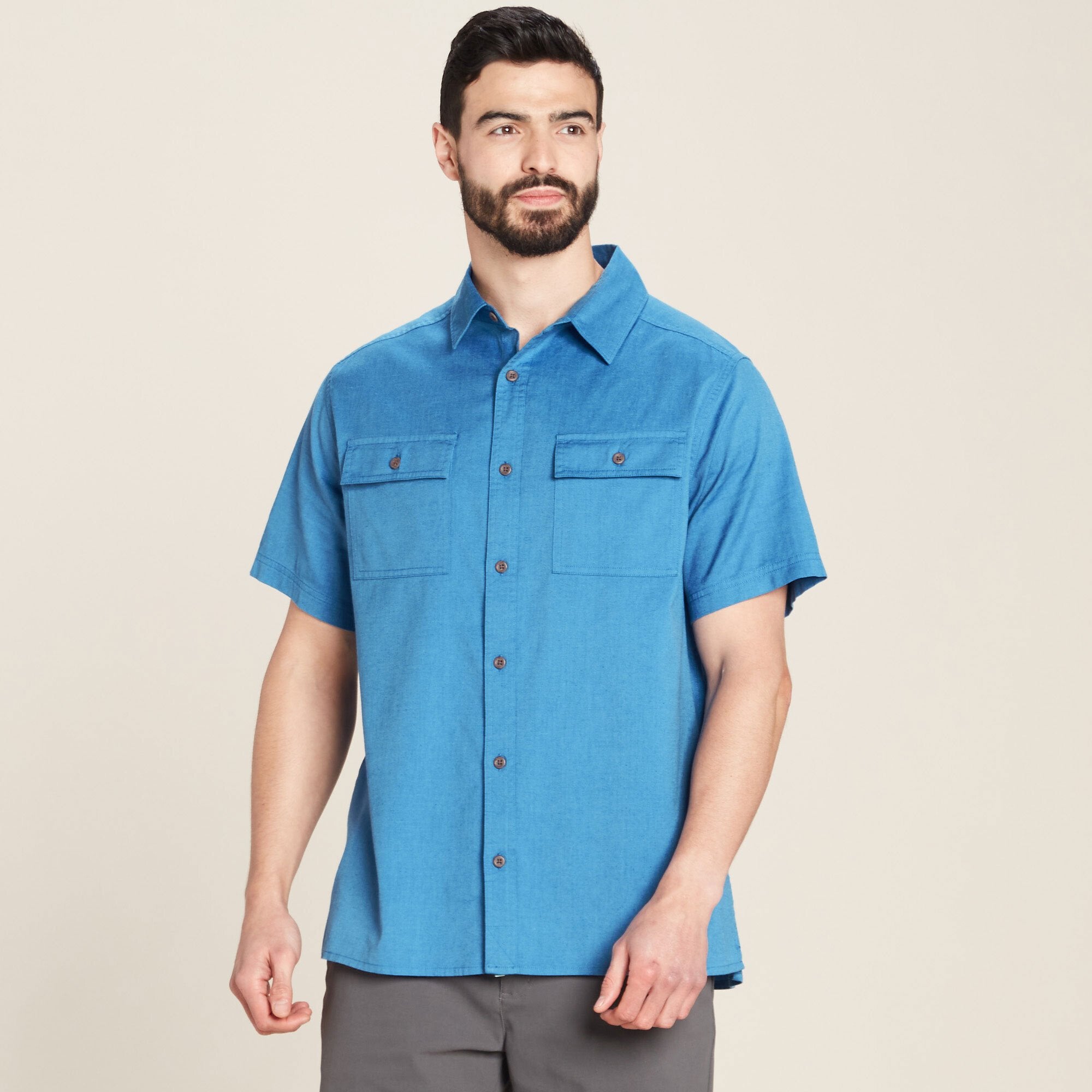 A man models a Sherpa Adventure Gear Tharu Journey Short Sleeve Shirt in Blue. The shirt features a structured collar and two chest pockets with button closures. The model is looking to the side, standing in a casual pose with his hands relaxed at his sides. He pairs the shirt with gray trousers, and the neutral background highlights the vibrant blue tone of the fabric.